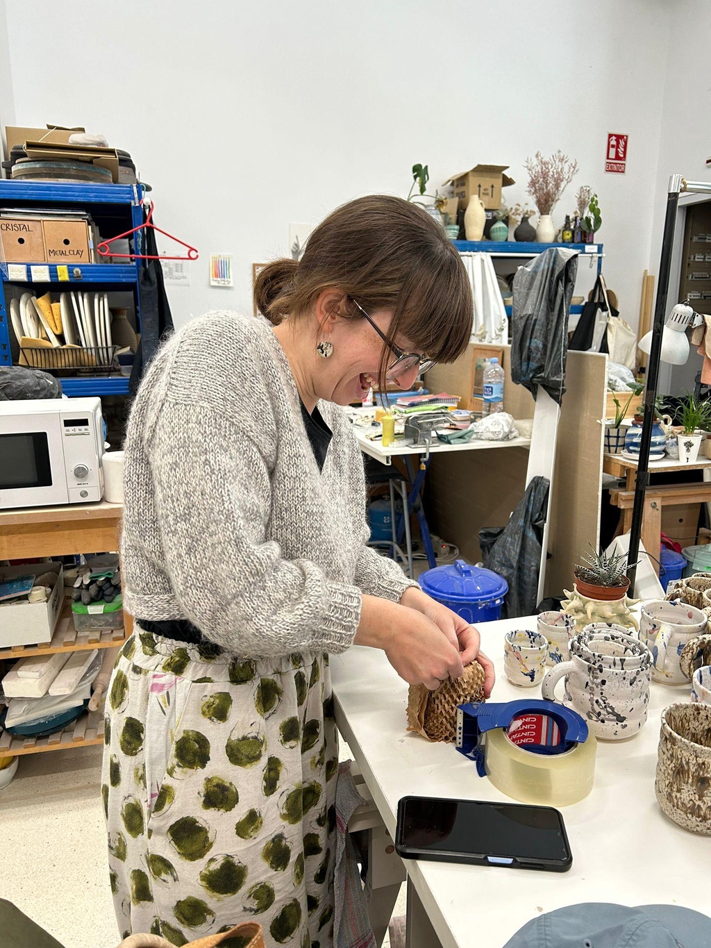 The picture shows Mirjam in the ceramic studio, laughing while wrapping up a cup in protective paper bubble wrap. In the foreground you can see her work table covered in ceramic bubble cups in different sizes. In the background, you can see her colleagues work tables and shelves.