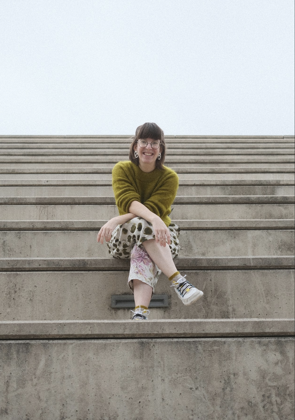 Mirjam is a brown haired caucasian woman in her thirties. She is sitting cross legged and smiling on concrete stairs, wearing a luminous, green, handknit jumper and loose fitting linen pants with a green, pink and white pattern. 