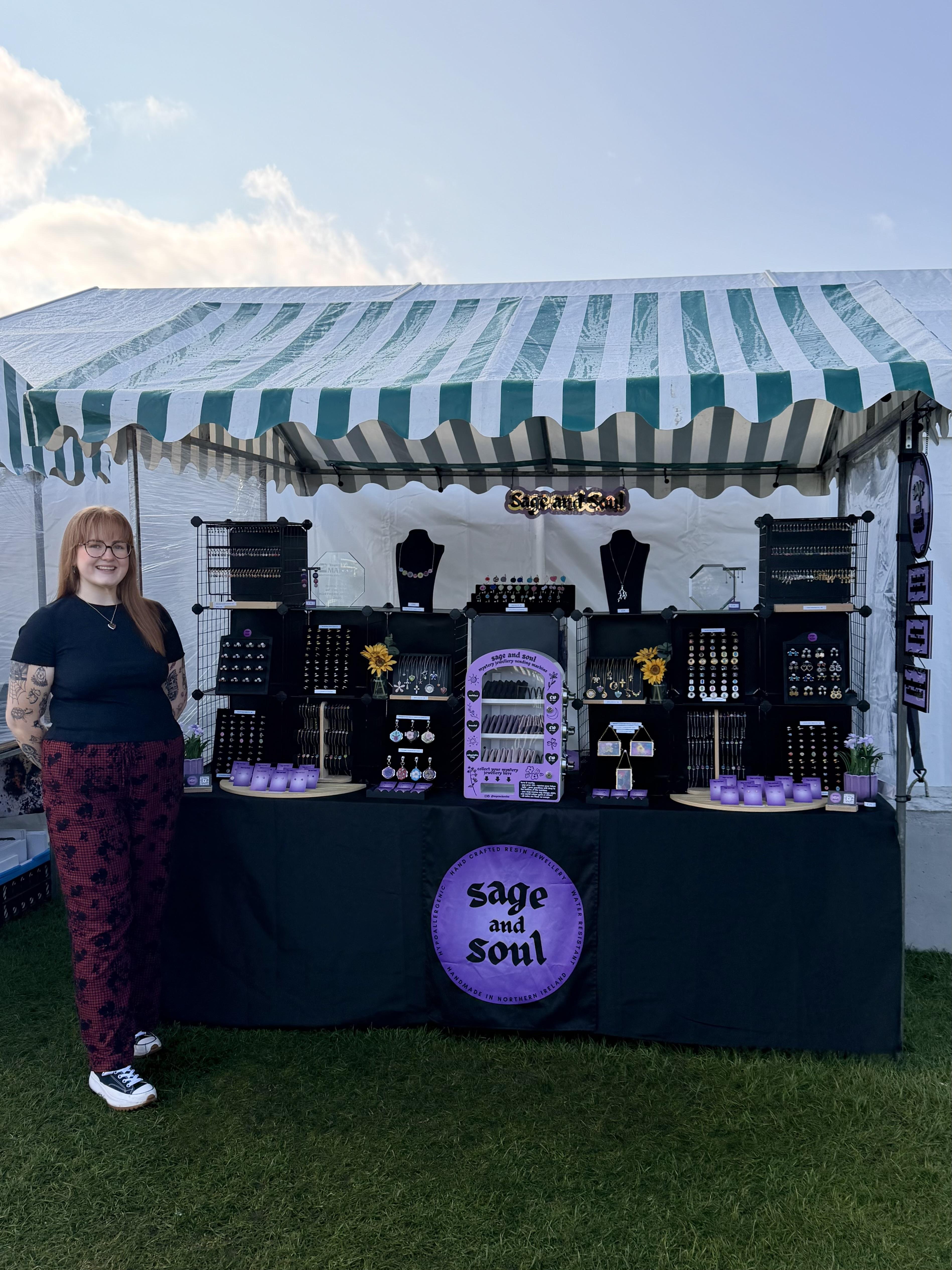 Amy, the owner of Sage and Soul, standing for a photograph, standing at her market stall, wearing a polka dot blouse and skirt.