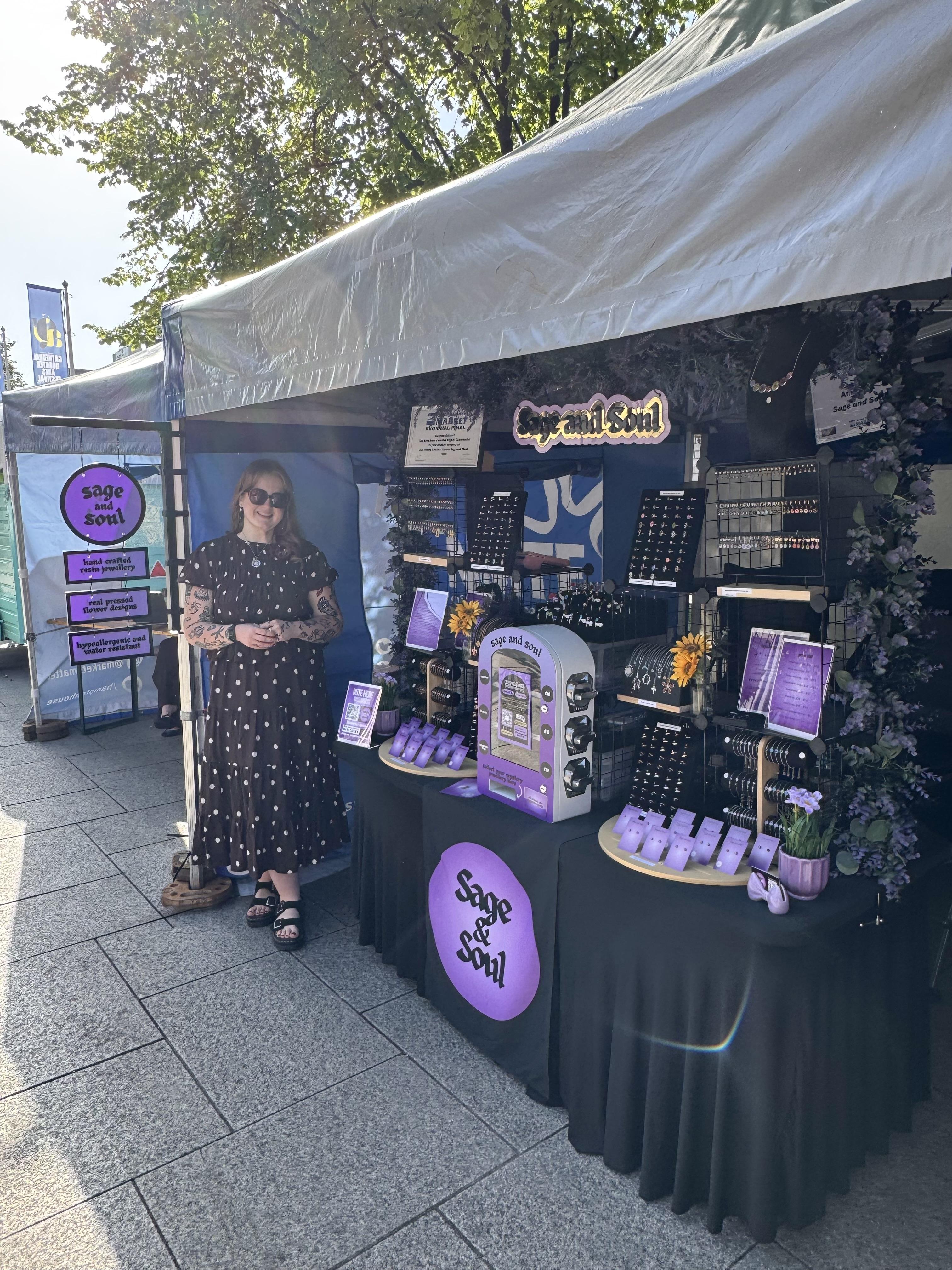 Amy, the owner of Sage and Soul, standing for a photograph, standing at her market stall, wearing a polka dot blouse and skirt.