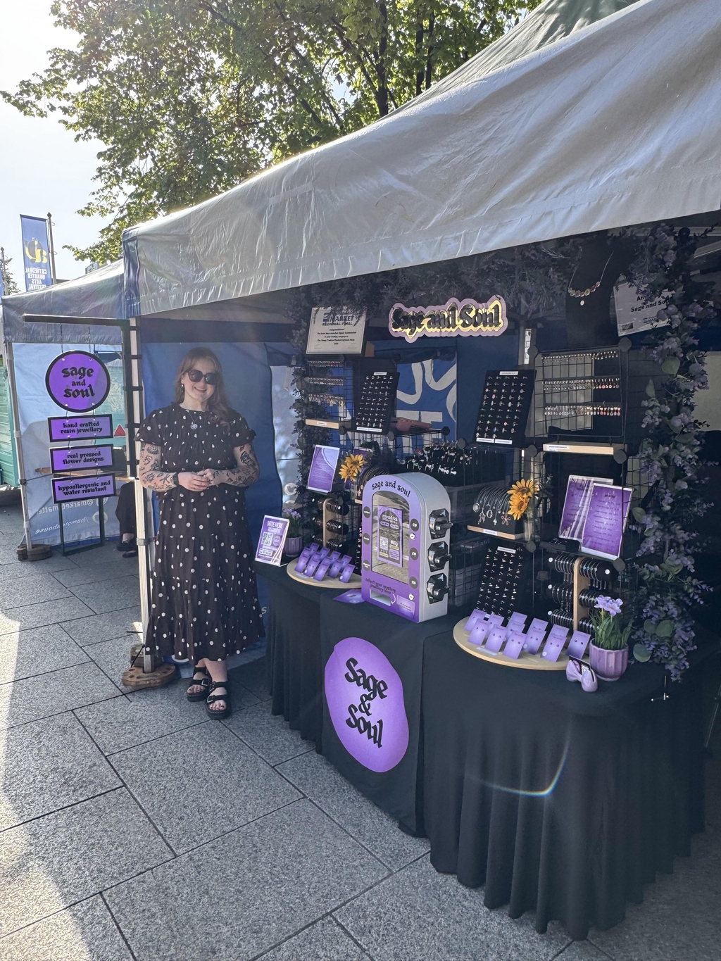 Amy, the owner of Sage and Soul, standing for a photograph, standing at her market stall, wearing a polka dot blouse and skirt.