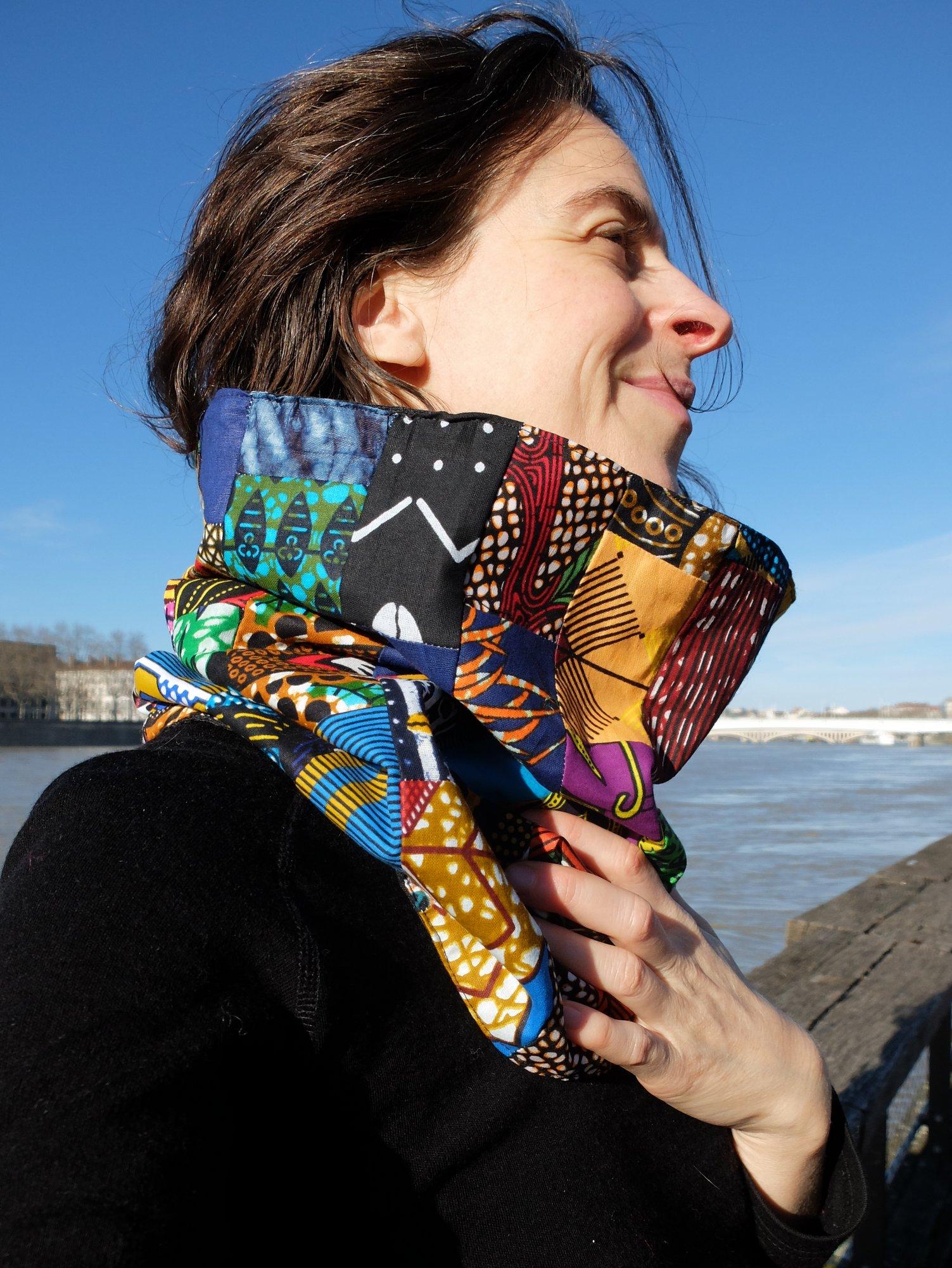 Cache-cou en patchwork wax et doublé de laine. Pièce unique, confection artisanale et éthique. Porté par une jeune femme souriant dans un cadre urbain devant un fleuve.