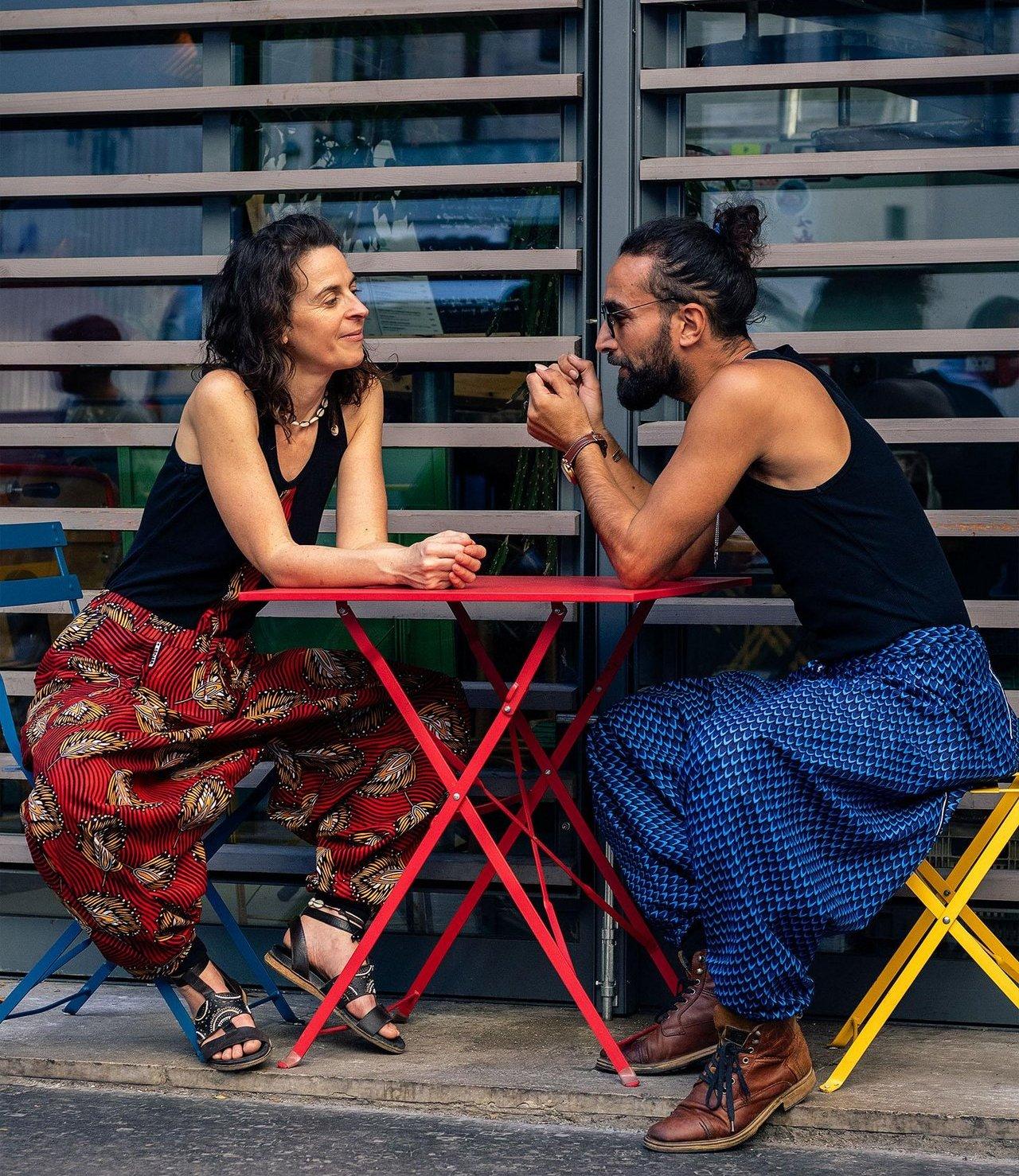 Pantalons sarouels en coton wax coloré, coupe ample et unisexe. Porté par un couple attablé en terrasse en été.