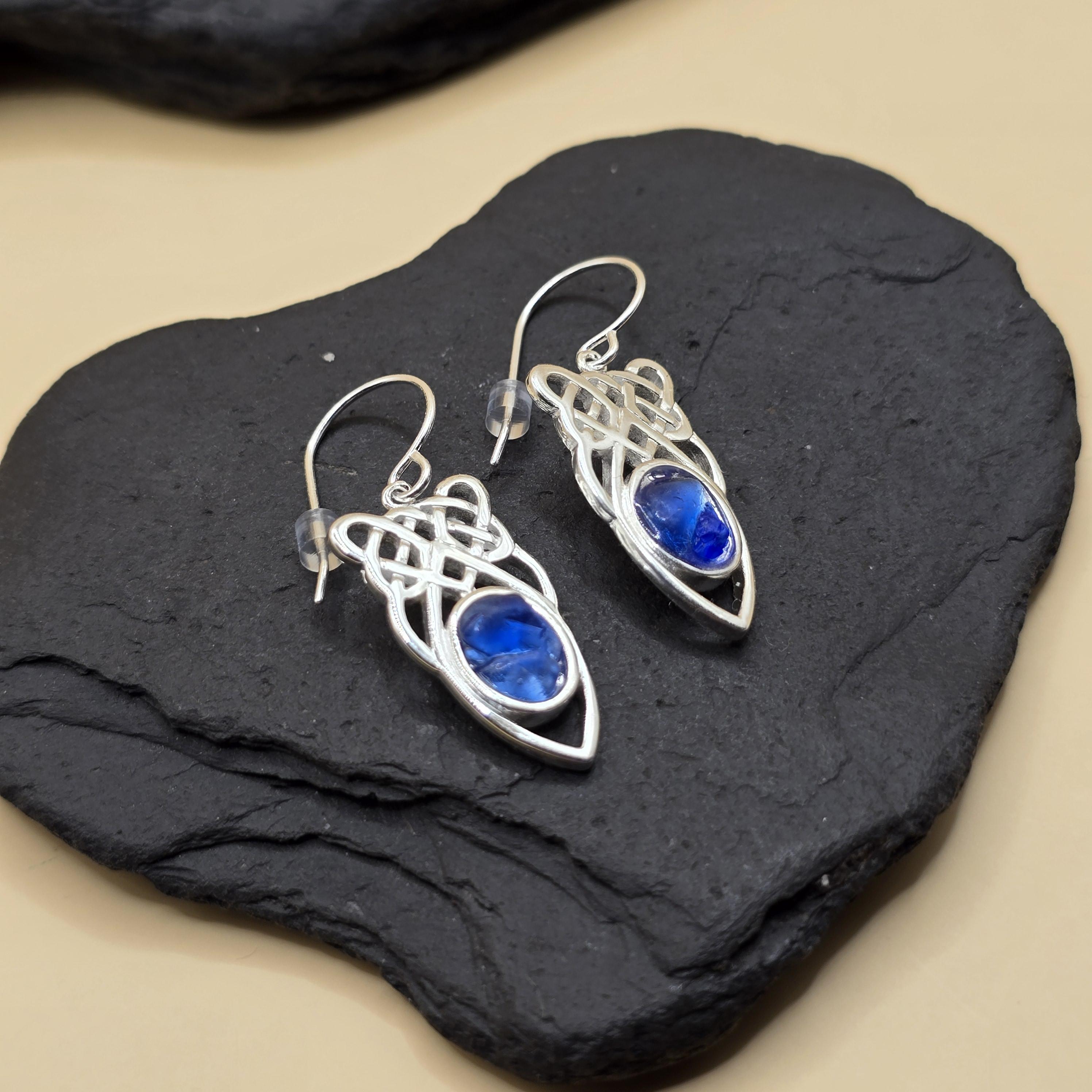 Handcrafted earrings featuring deep blue Scottish seaglass from Berwickshire beaches, set in silver-toned metal with an engraved floral and wave motif. The teardrop-shaped seaglass pieces are framed by a crescent-shaped base, combining natural coastal elements with intricate design. The earrings hang from simple silver hooks, offering a unique blend of beachcombed materials and artisan craftsmanship.