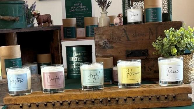 Five candles with colourful labels displayed among some vintage items and wooden boxes. Labels describe wellbeing themes: Restore, Comfort, Joyful, Revive and Peace