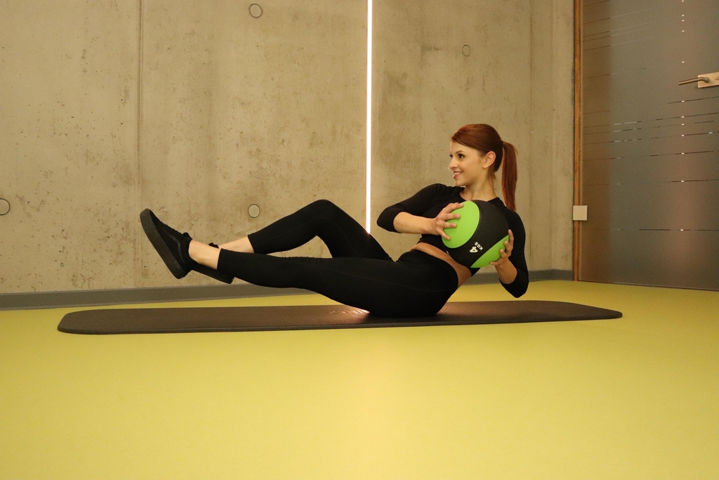 Bild von junger Frau die mit einem Medizinball auf der Matte die Übung Russian Twist ausführt. 
