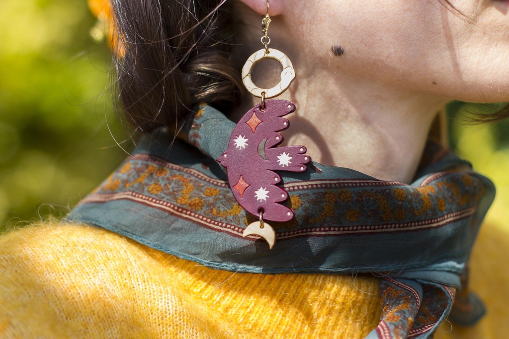 Boucle d'oreille portée par une personne qui porte un foulard vert foncé, les éléments de la boucle d'oreille sont : un serpent type ouroboros en bois, un oiseau en cuir, une lune en bois
