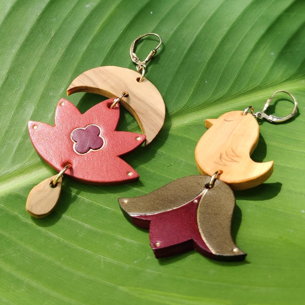 Une paire de boucles-d'oreilles  posée sur une feuille de bananier, les éléments des boucles sont en bois et en cuir, ils représentent les éléments suivants: oiseau, lune, fleur, goutte