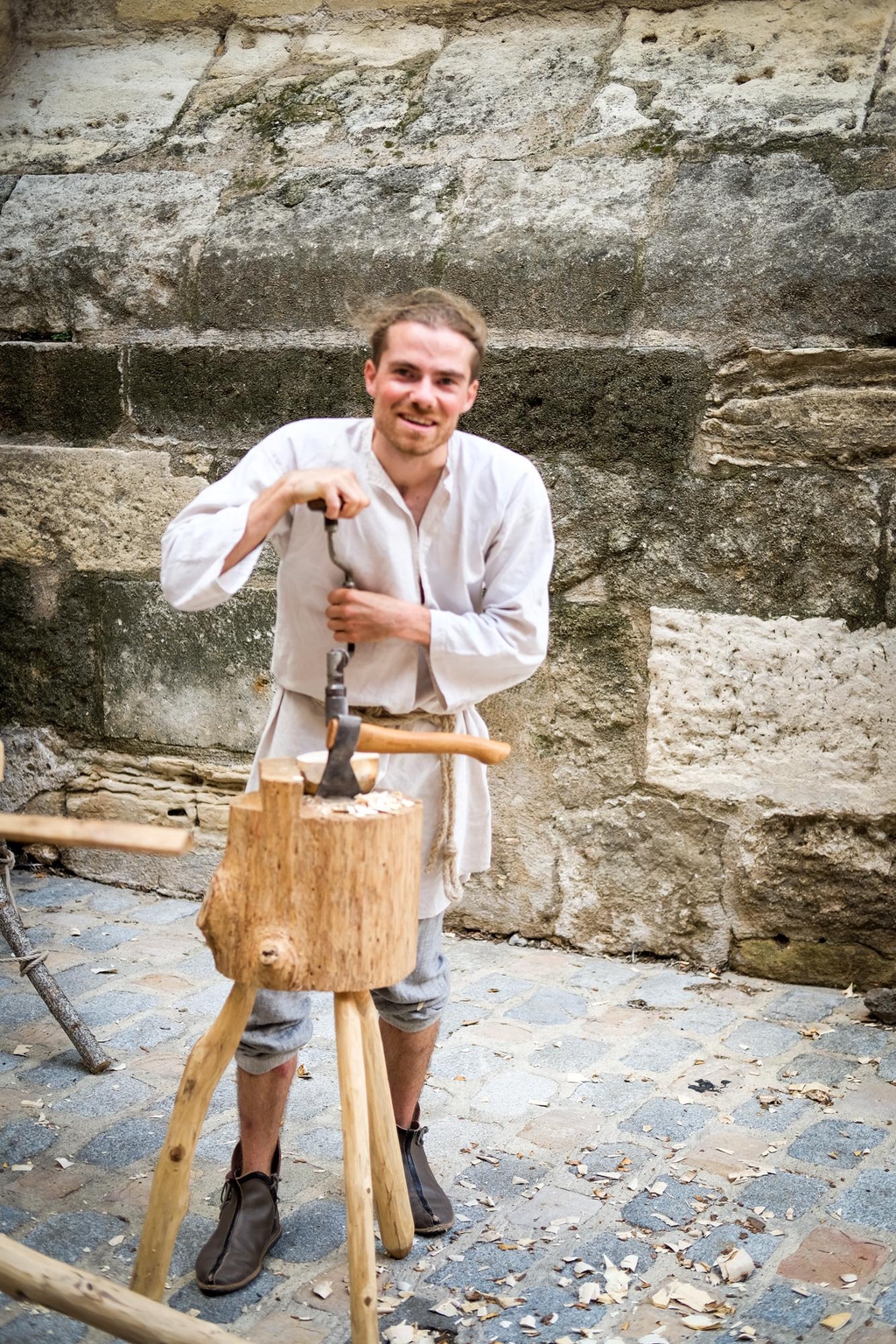 Un artisan perce un bol en bois avec une chignole, il travaille sur un billot en bois dans lequel est enfoncé une hache