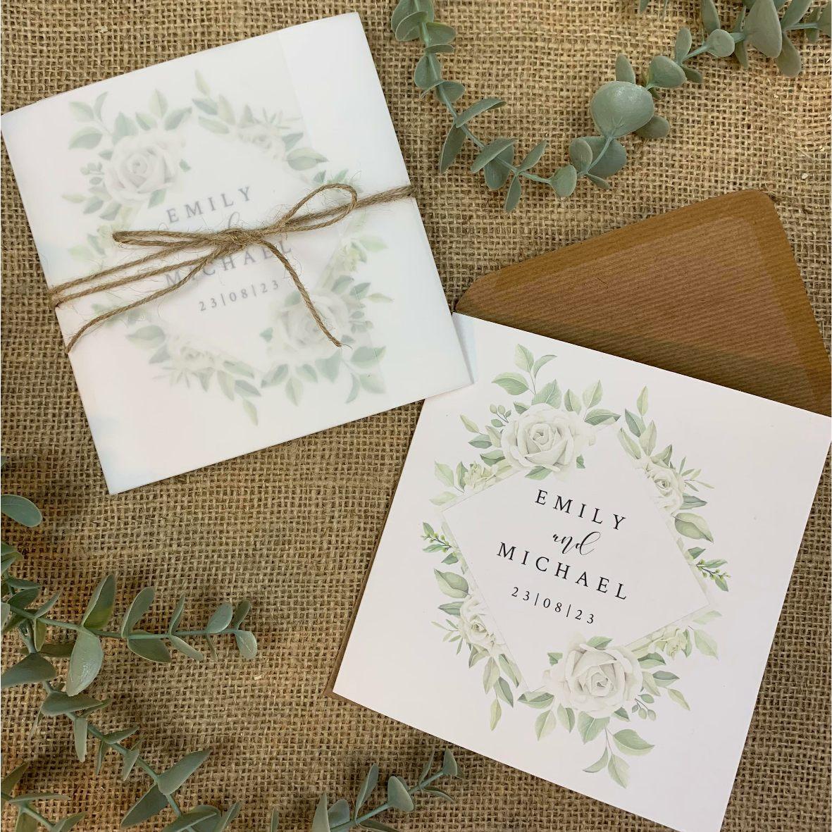 Image of white rose wedding invitations with brown kraft paper envelope. One of the invitations is wrapped in a vellum jacket and tied with rustic twine.