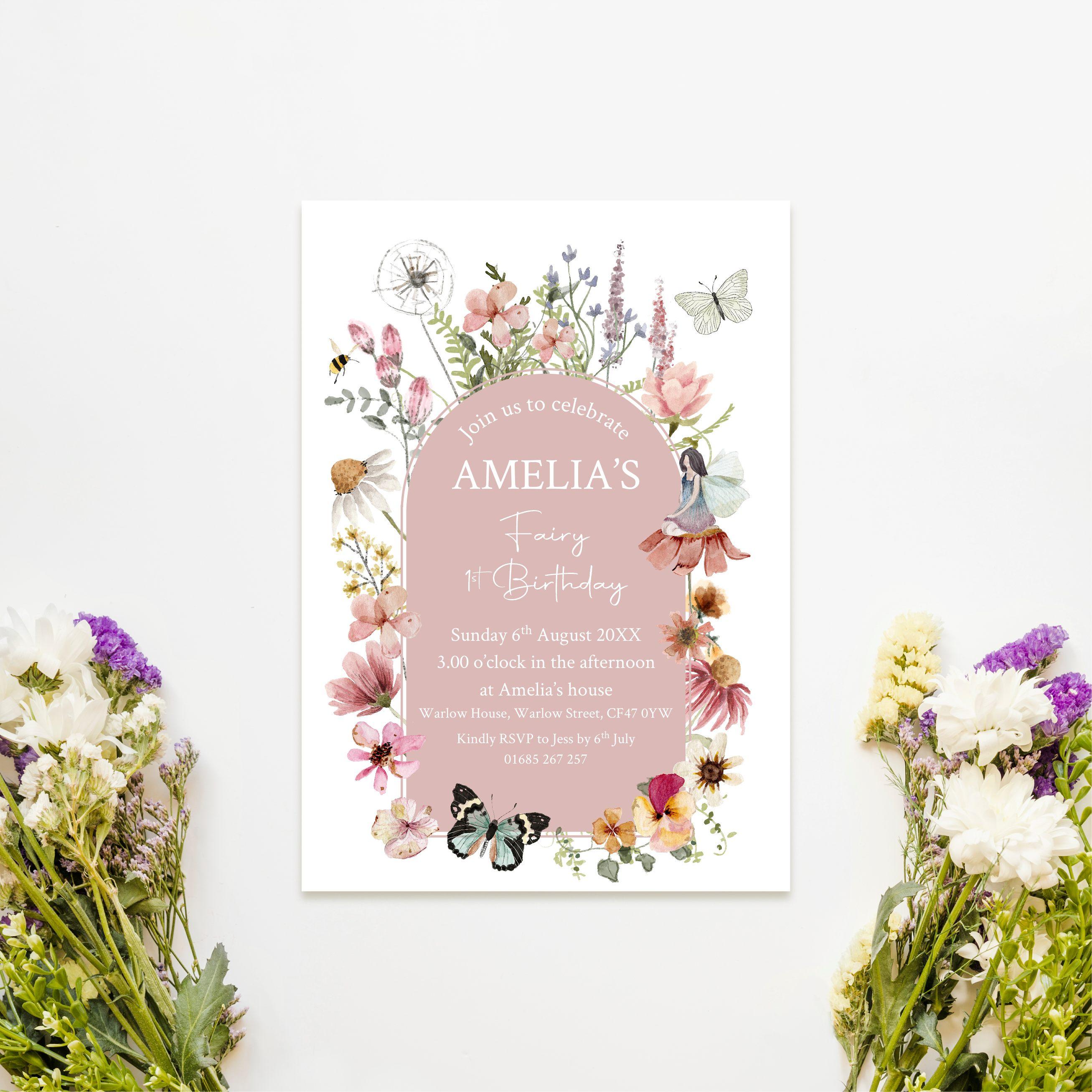 An image of an A6 postcard invitations for a first birthday party. The central design is blush pink with the wording in white. The central design is surrounded by wildflowers in shades of pink, yellow, white and green. There is a small fairy sat on a flower on the right hand side and butterflies & a bee also feature in the design.