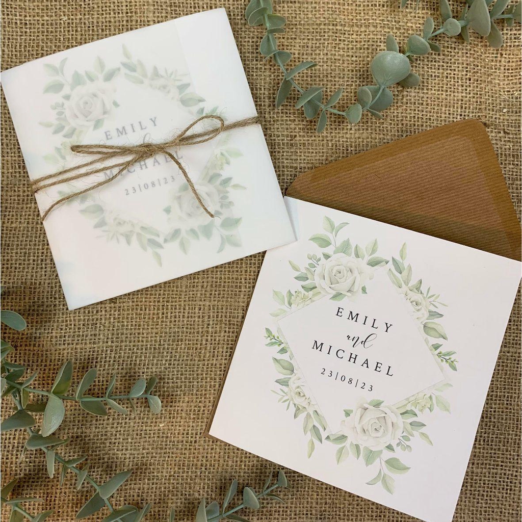 Image of white rose wedding invitations with brown kraft paper envelope.  One of the invitations is wrapped in a vellum jacket and tied with rustic twine.