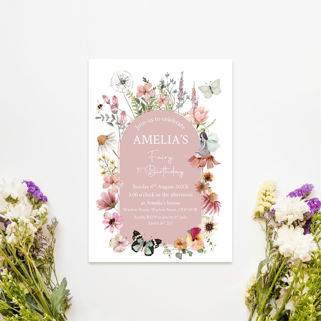 An image of an A6 postcard invitations for a first birthday party. The central design is blush pink with the wording in white. The central design is surrounded by wildflowers in shades of pink, yellow, white and green.  There is a small fairy sat on a flower on the right hand side and butterflies & a bee also feature in the design.