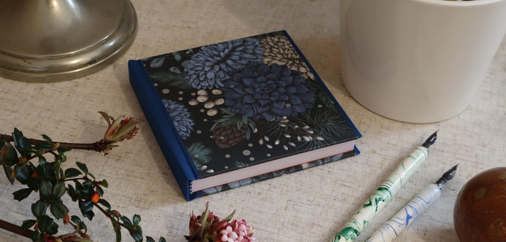 A traditionally handmade notebook or journal in blue cloth and a winterly pattern of flowers, pinecones and sprinkles of snow.