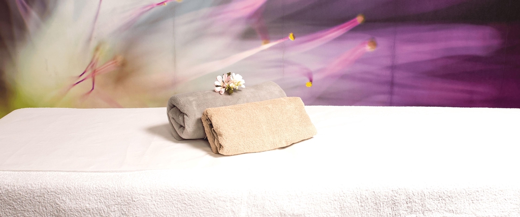 Cabina per massaggi, con lettino pronto all’uso in primo piano e sfondo floreale viola.
