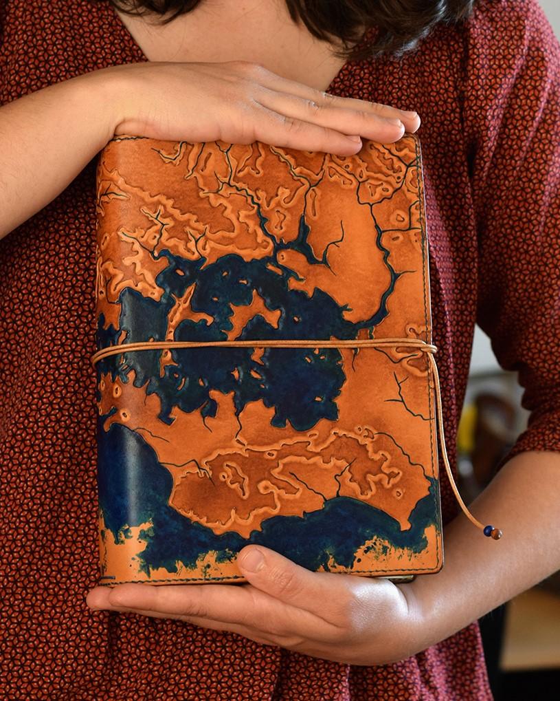 Photo d'un couvre-carnets en cuir de la marque Helix échappée cuir. Créé sur-mesure, il représente une carte topographique du golfe du Morbihan (Bretagne) mise en relief par la technique du repoussage.