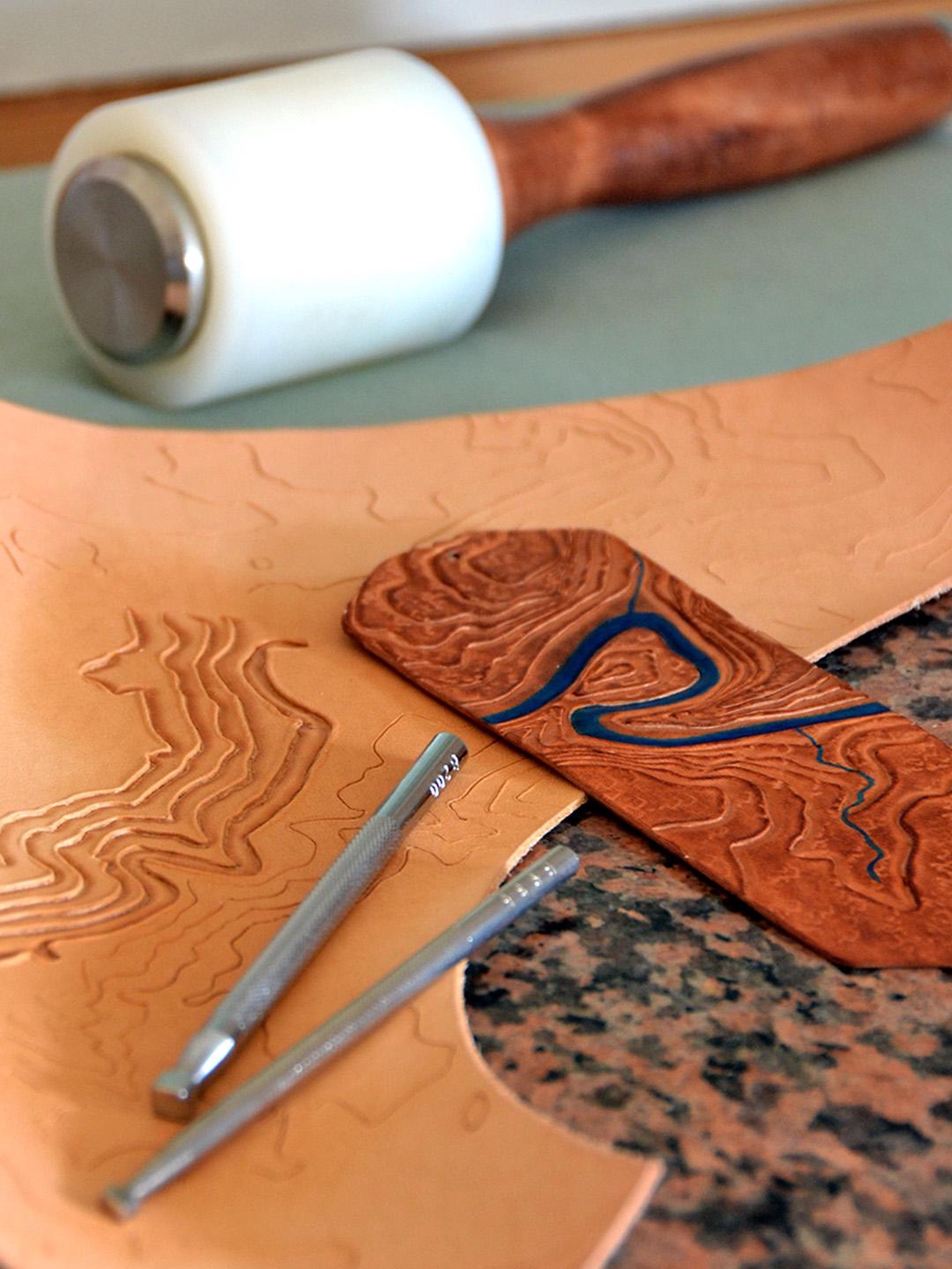 Photo d'une pièce de cuir tanné végétal en cours de repoussage (=mise en relief) d'un motif topographique. Des matoirs (outils utilisés pour le repoussage) et un marque-pages terminé sont posés dessus.