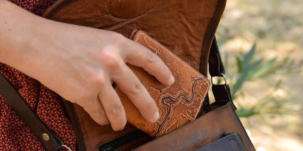 Photo d'une main qui glisse un porte-cartes de la marque Helix échappée cuir dans un sac. Une carte topographique mise en relief par la technique du repoussage orne l'extérieur de ce porte-cartes.