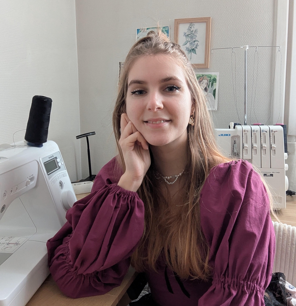 Femme blonde souriante avec un haut violet posant à côté d'une machine à coudre