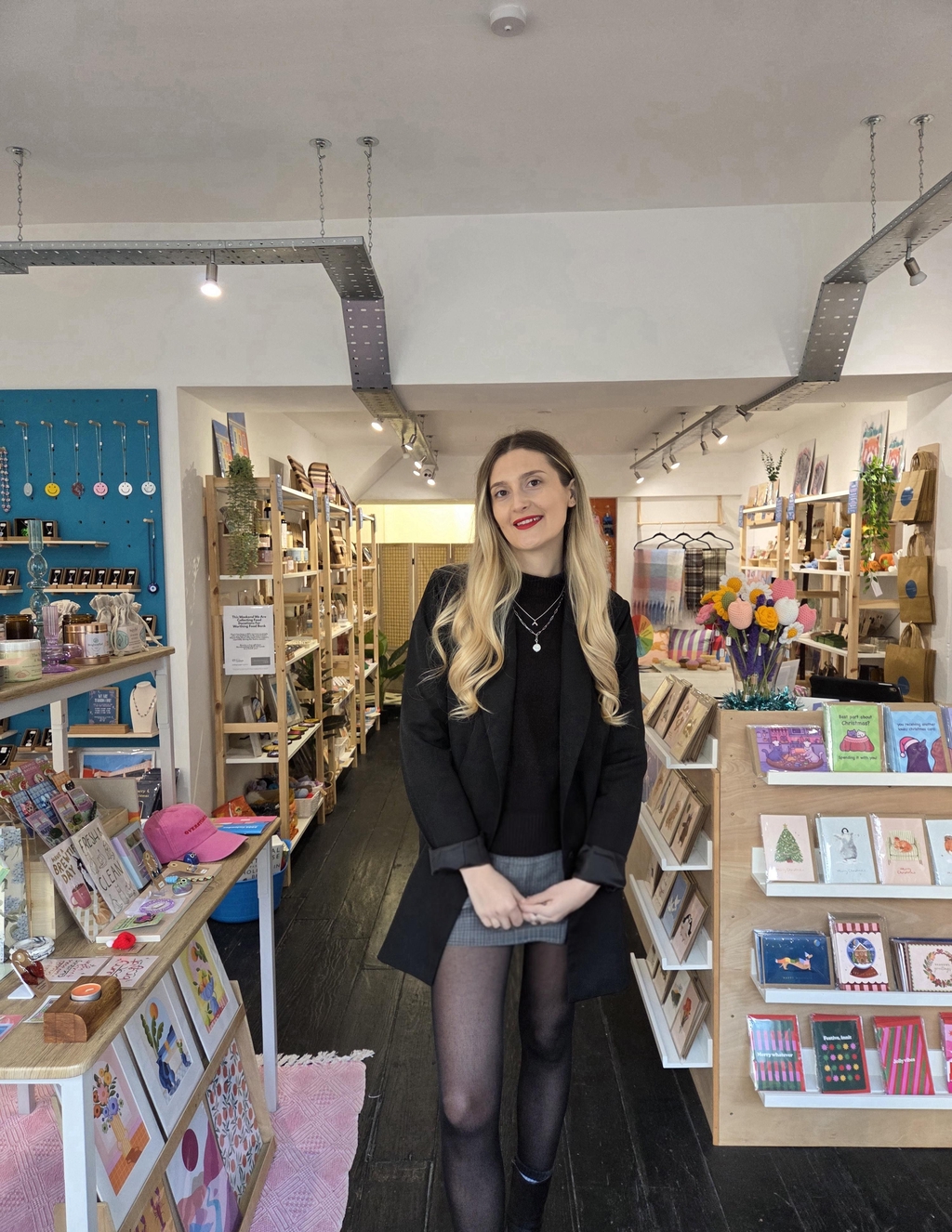 A young mid 20s year old female wearing a black jumper, black blazer and a grey skirt with tights. Wearing bold red lipstick and long blonde hair in a bouncy blowdry curled style. She wears two silver necklaces one with a cubic zirconia rainbow charm and the other with a coin pearl. She is smiling and happy as she stands in the middle of her shop, surrounded by handmade goodies from UK independent makers.