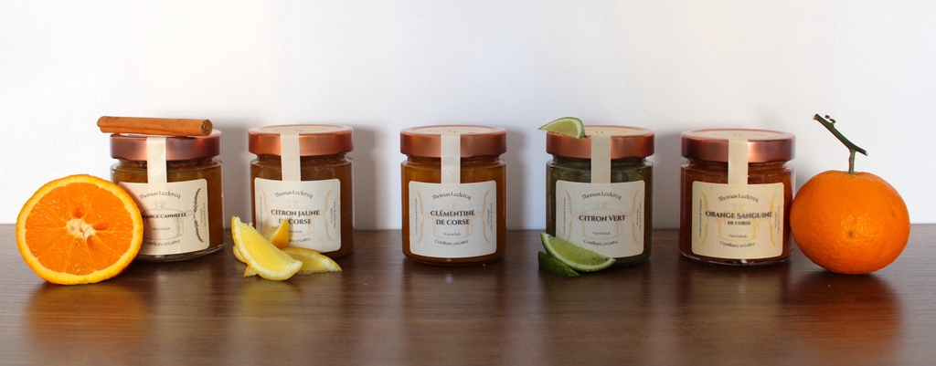 5 pots en verre avec couvercle en cuivre haut de gamme contenant une variété diverses de marmelade d'agrumes : Orange/cannelle, Citron jaune de Corse, Clémentine de Corse, Citron Vert, Orange sanguine de Corse. Des tranches d'agrumes et d'épices utilisés dans les marmelades sont utilisés pour compléter et décorer le scène.