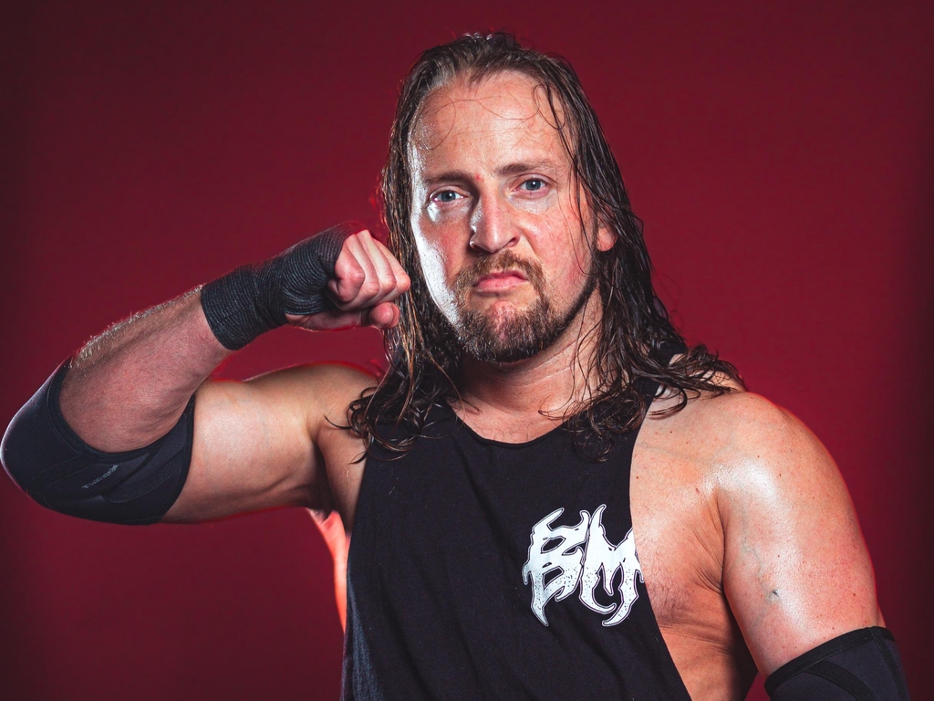 Brad Matthews stand in his black vest in front of a dark red background.