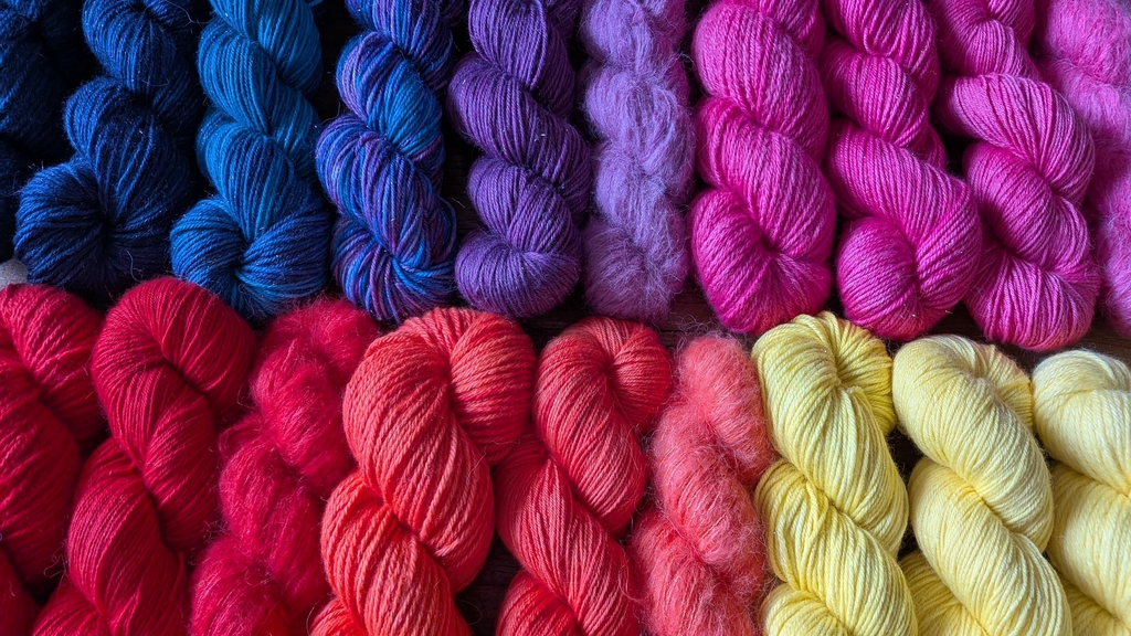 Assortiment coloré de fils à tricoter Cosmic Yarns. La photo montre plusieurs écheveaux de laines teintes à la main de différentes compositions (Mérinos Superwash, Mérinos non-Superwash, Baby Suri Alpaga et Soie) et grosseurs (lace, fingering). Les coloris variés incluent des bleus cosmiques intenses, des roses nébuleuses, des oranges, des jaunes, des violets, et des rouges vifs, les unes à côtés des autres comme un dégradé de couleurs.