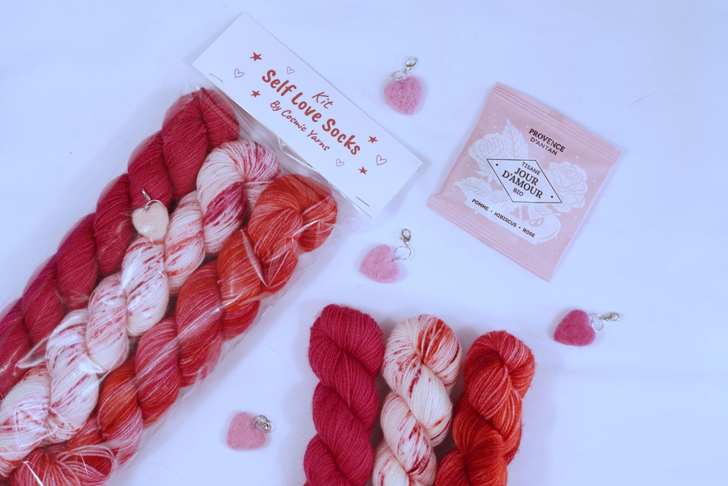 Kit self love socks. Sachet de 3 écheveaux de 50g/212m dans 3 coloris (rose foncé, blanc à speckles rose et rouge, et rouge/rose mélangés), marqueur de mailles feutrés à la main en forme de coeur et sachet de tisane à la pomme, hibiscus et rose