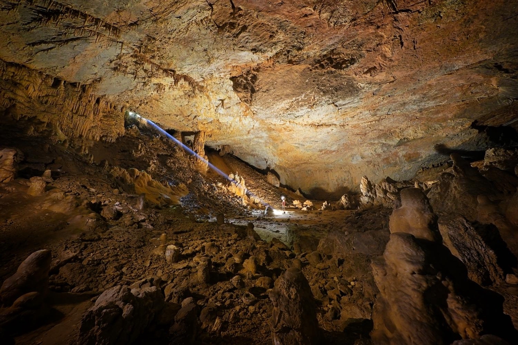 A man stands in a large cave chamber with a beam of sunlight from the entrance illuminating him. 