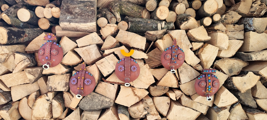 As a backdrop, a stacked pile of wooden logs is arranged neatly, showcasing various sizes and textures of the wood. In the foreground are 5 ornate, African-inspired ceramic masks. 