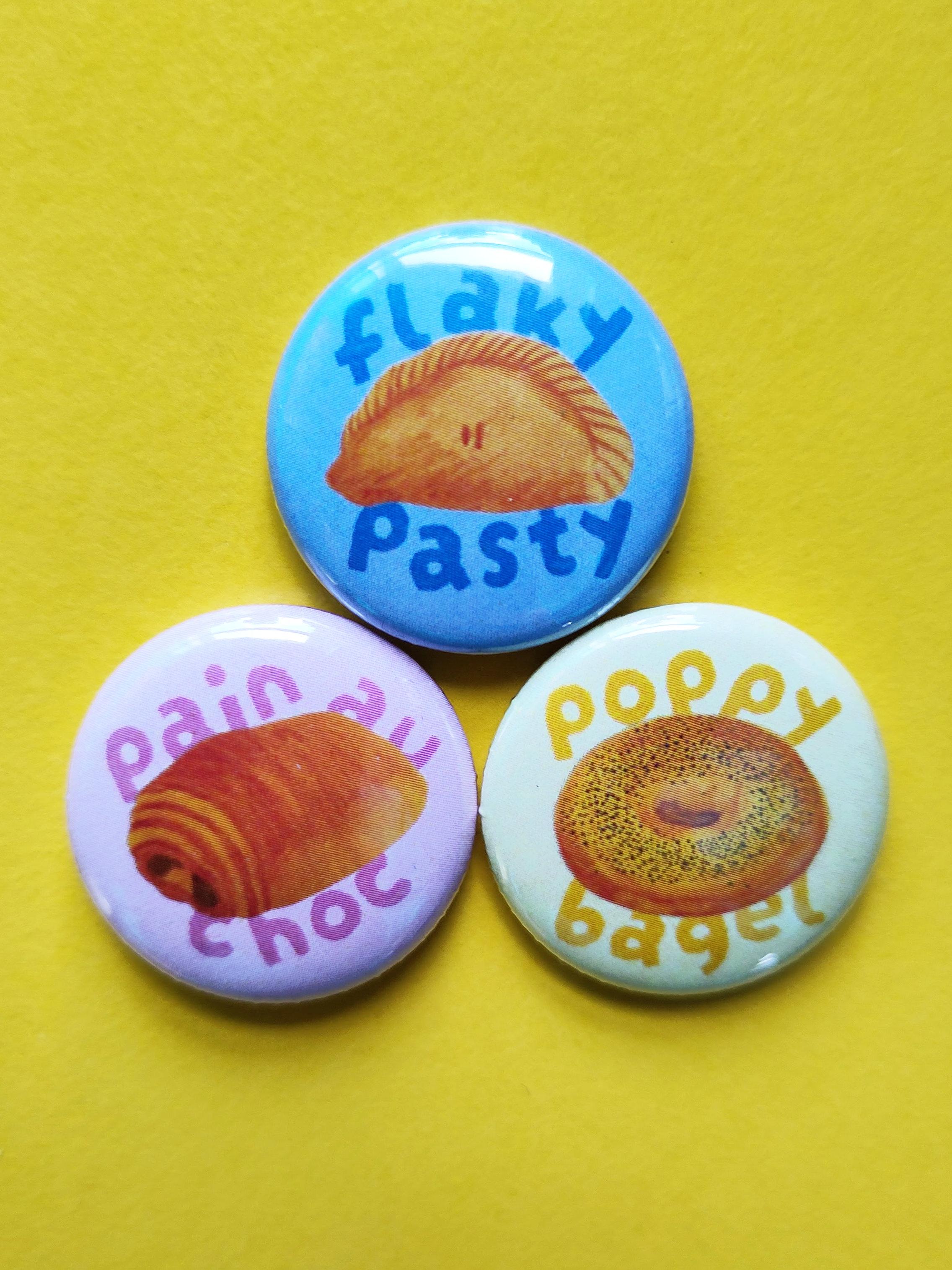 Three bakery themed pin badges with cute mini watercolours and chunky text. There's a blue badge with a pasty, a yellow badge with a bagel and a pink badge with a pain au chocolat.