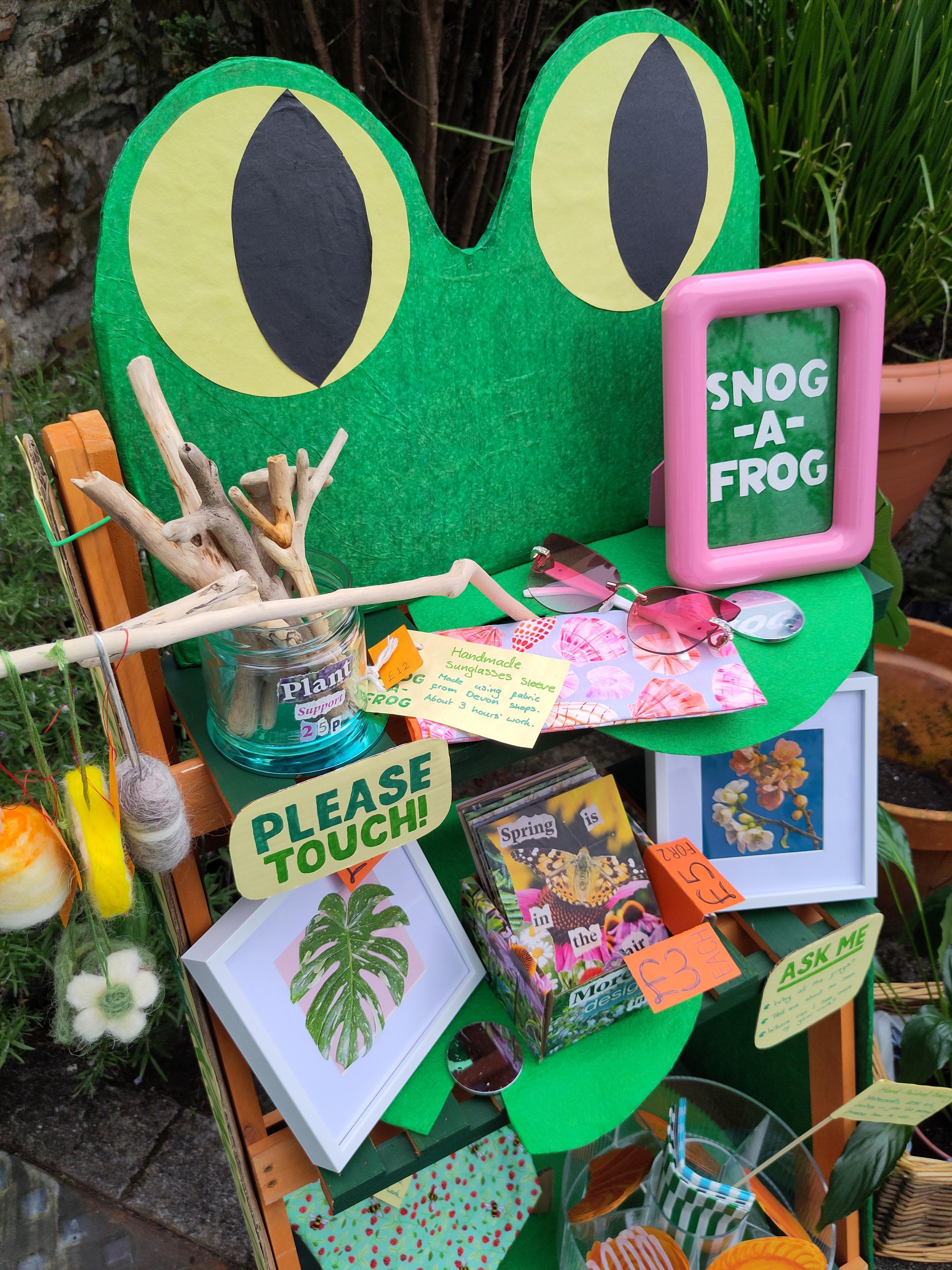 Large cardboard frog eyes on top of a folding shelf. The shelf is decked with handmade wares, including a sewn sunglasses sleeve in pinks and oranges, handmade cards, framed art and driftwood plant supports. Everything is colourful and friendly.