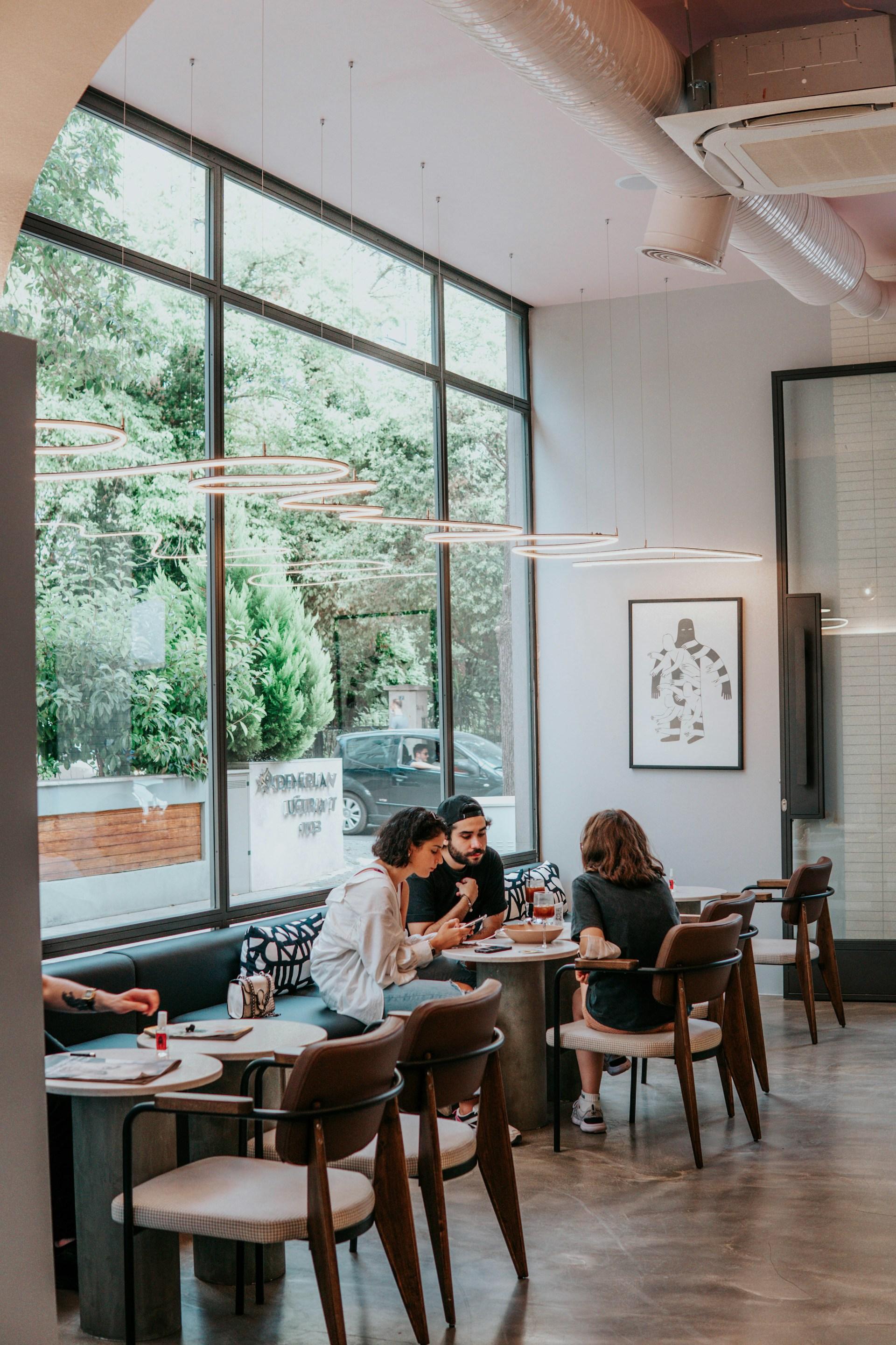 Three people chatting at a cafe table a distance away. Behind them are floor to ceiling windows that show a leafy street outside. Photo by Ceyda Çiftci.