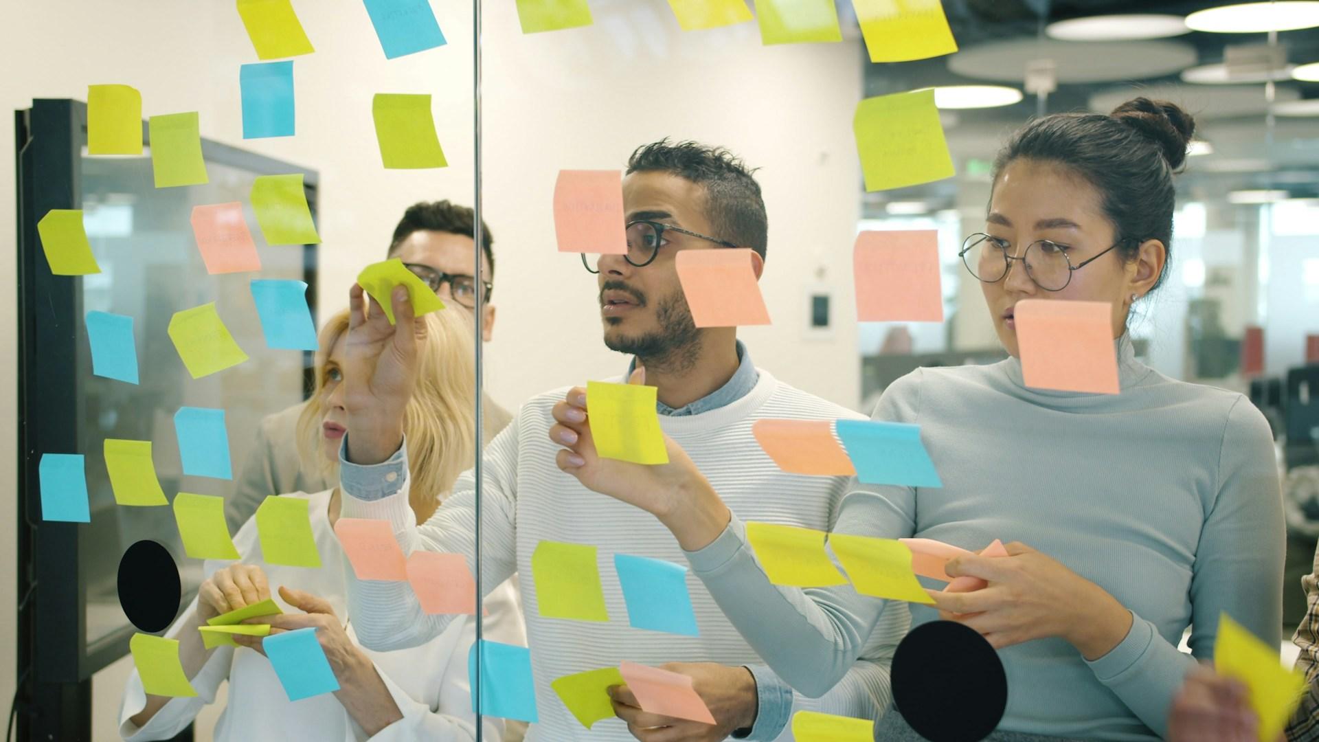 Four people on the other side of a large pane of glass. They're sticking different coloured sticky notes to the glass as part of a brainstorming meeting. Photo by
Vitaly Gariev.