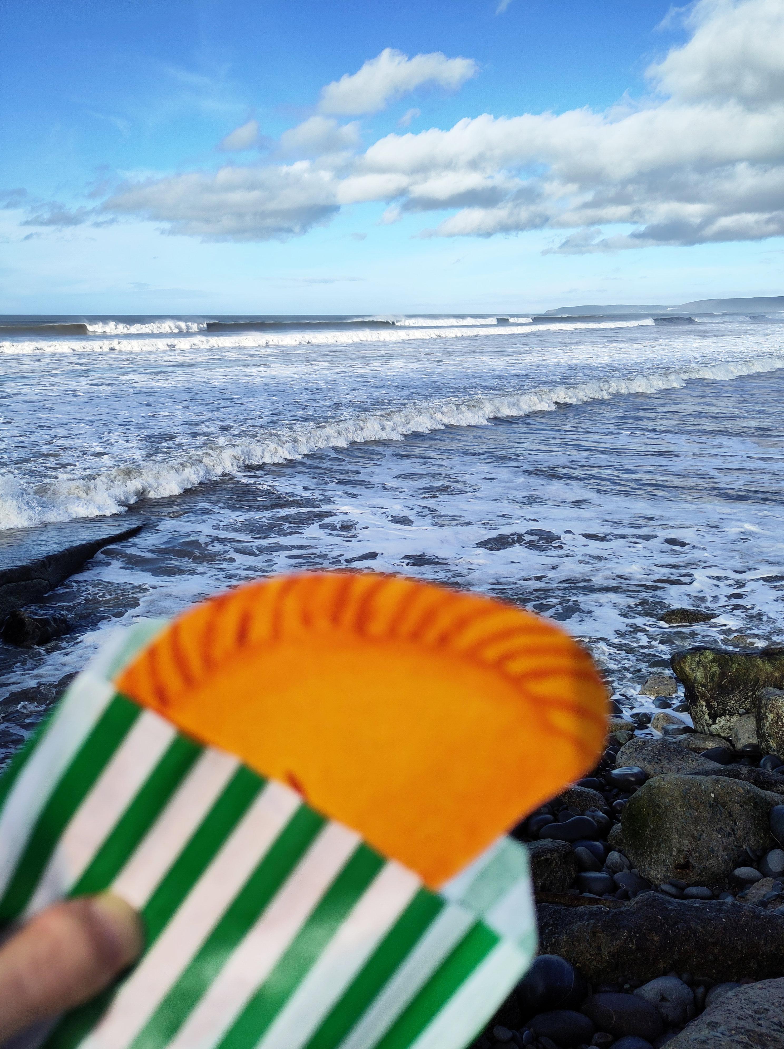 A blurry close-up of a watercolour pasty in a green and white stripy paper bag. In the background are crashing waves, in focus.