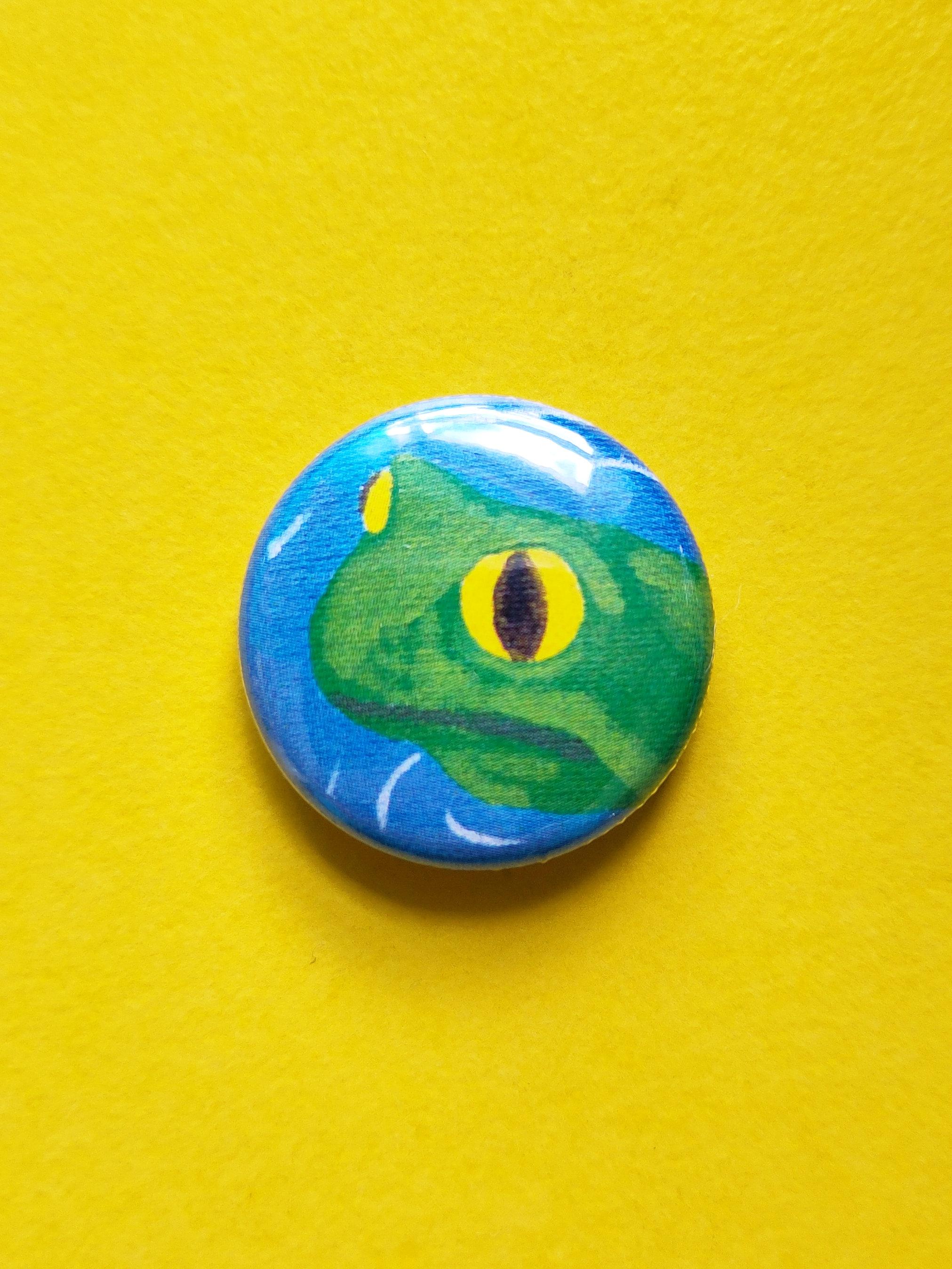 A pin badge of a bewildered, vacant-eyed frog sitting in a pond. The colours are flat and stylised like pop art. The badge is resting on a bright yellow background.