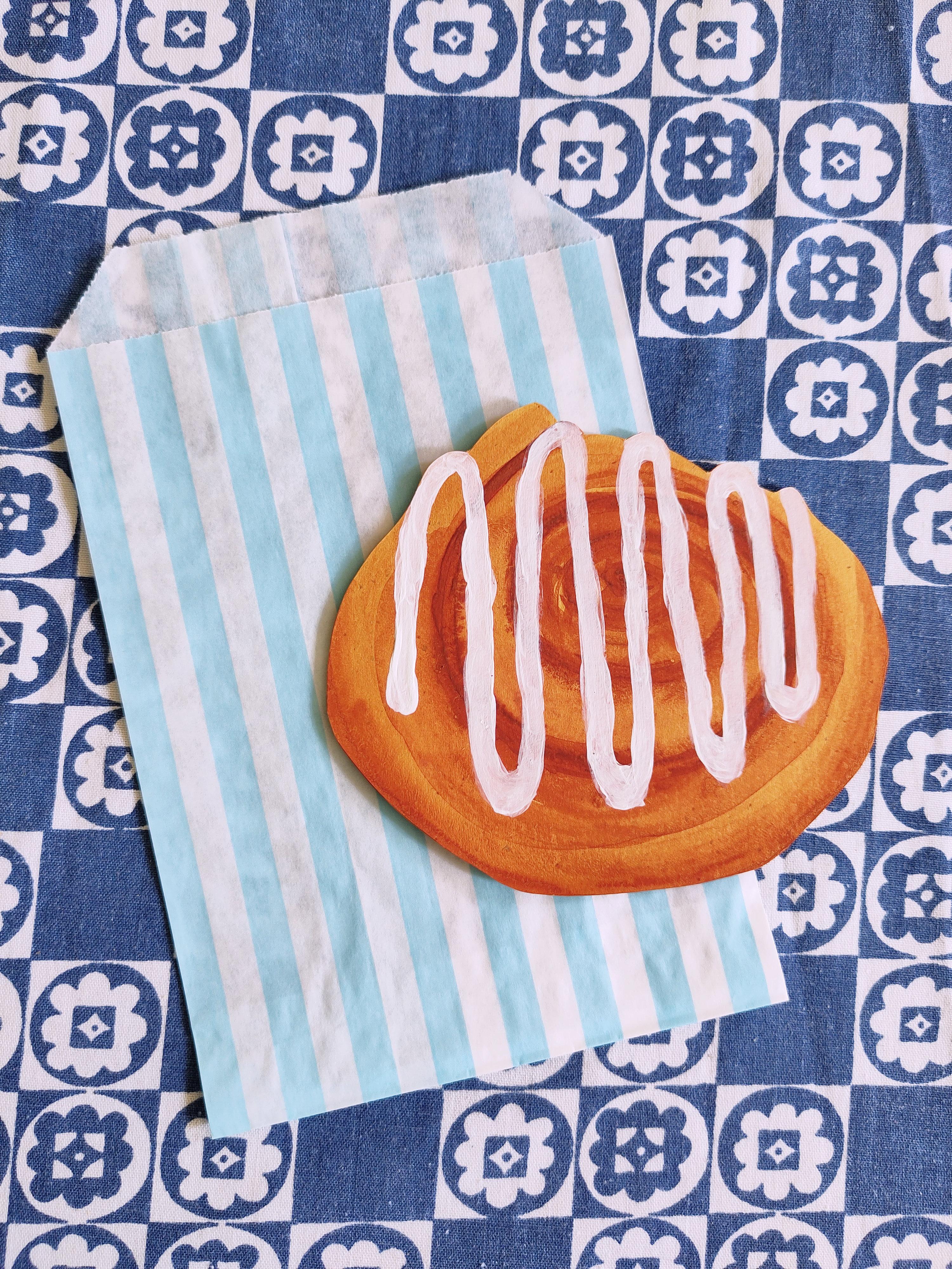 A cut-out watercolour of a cinnamon roll with a streak of acrylic icing. It's paired with a light blue and white stripy paper bag. It's resting on a patterned dark blue and white tablecloth.