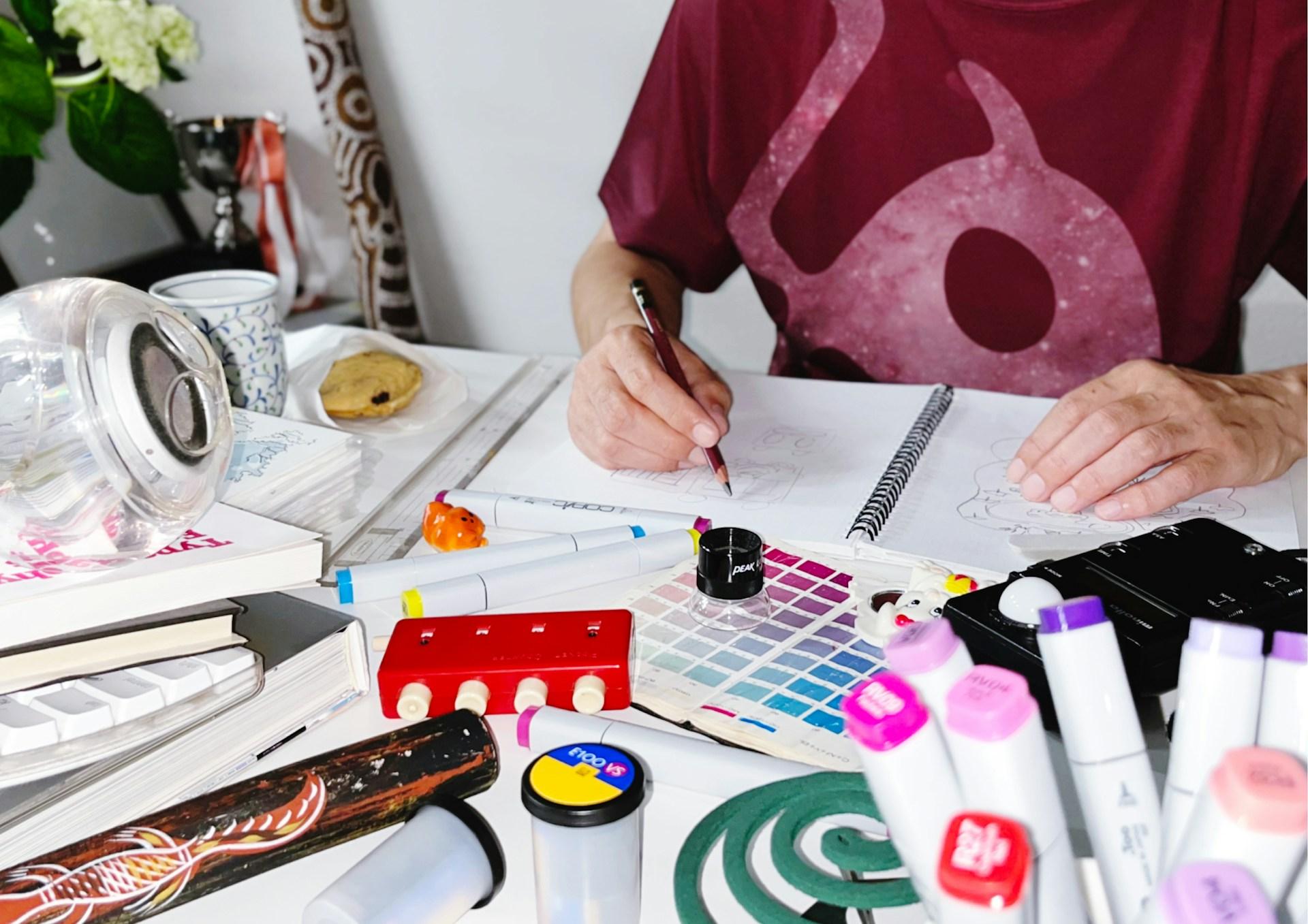 A desk covered in sketchbooks, pens and colour charts. There's the middle part of someone's body sitting behind the table. They're drawing in the sketchpad. Photo by K Nagahashi.
