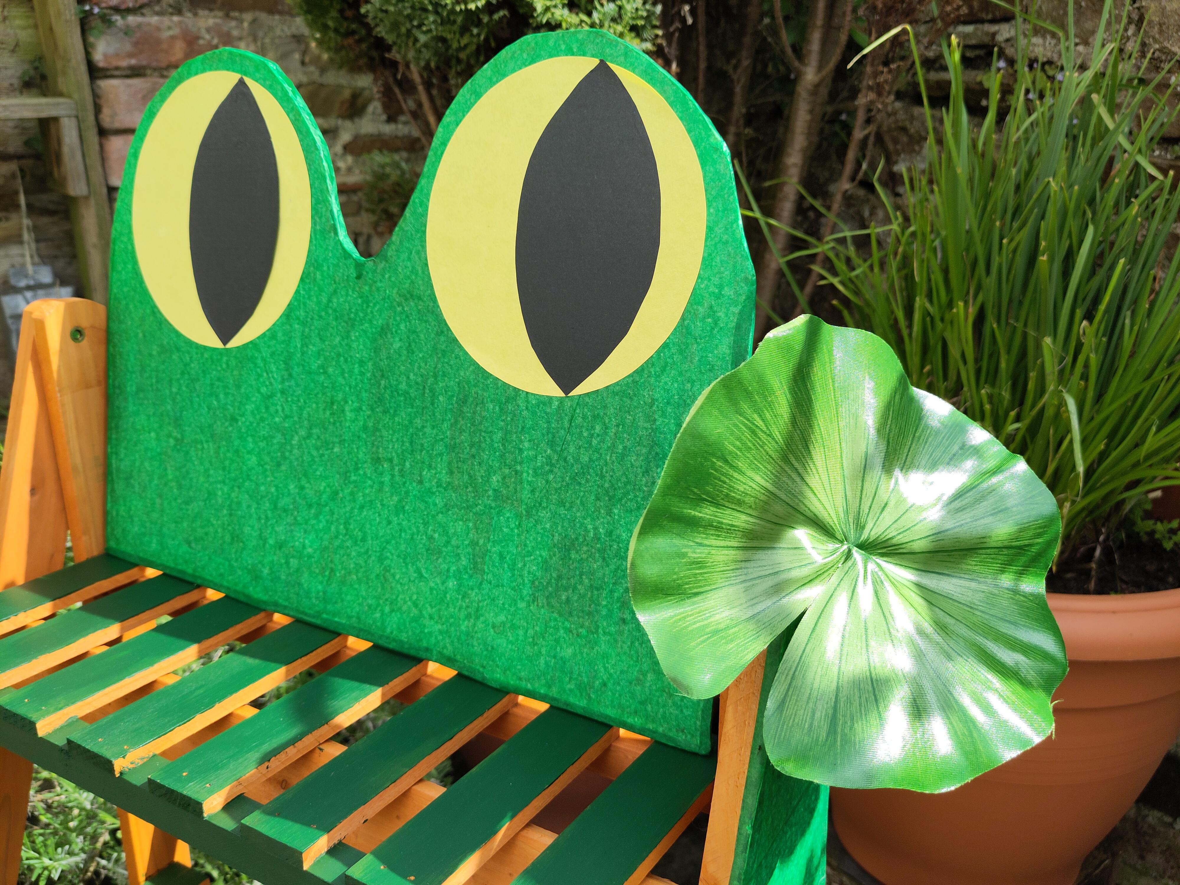 A pair of large, cardboard frog eyes in green and yellow. They're sitting on top of a folding shelf in a garden. There's a silky lilypad attached to the corner.