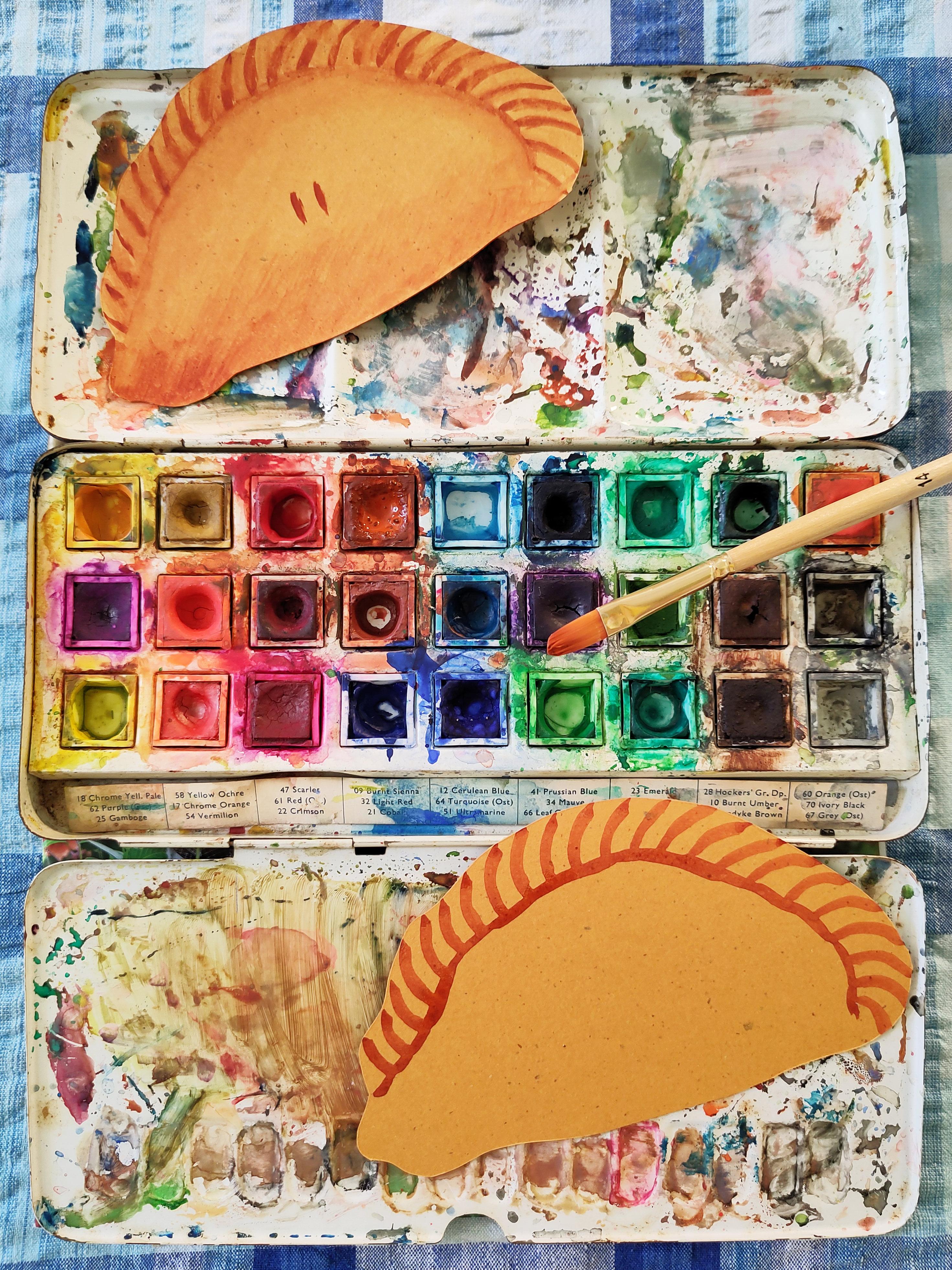 A messy tray of watercolours. Two pasty illustrations are resting on top, one finished, the other in progress with no shading.