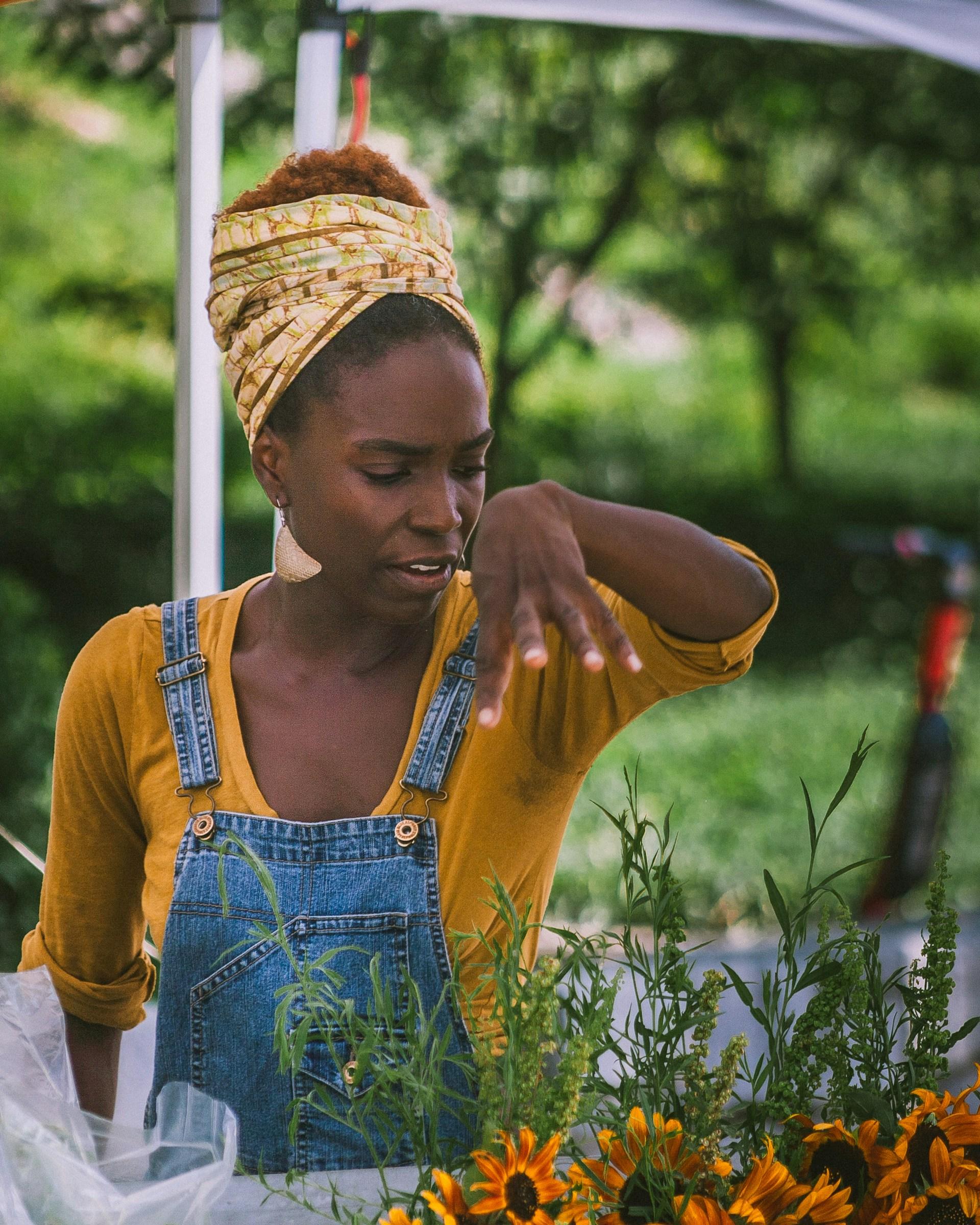 A black woman wearing dungarees. She's standing in a green park and gesturing at some herbs. Photo by Bryan Turner.