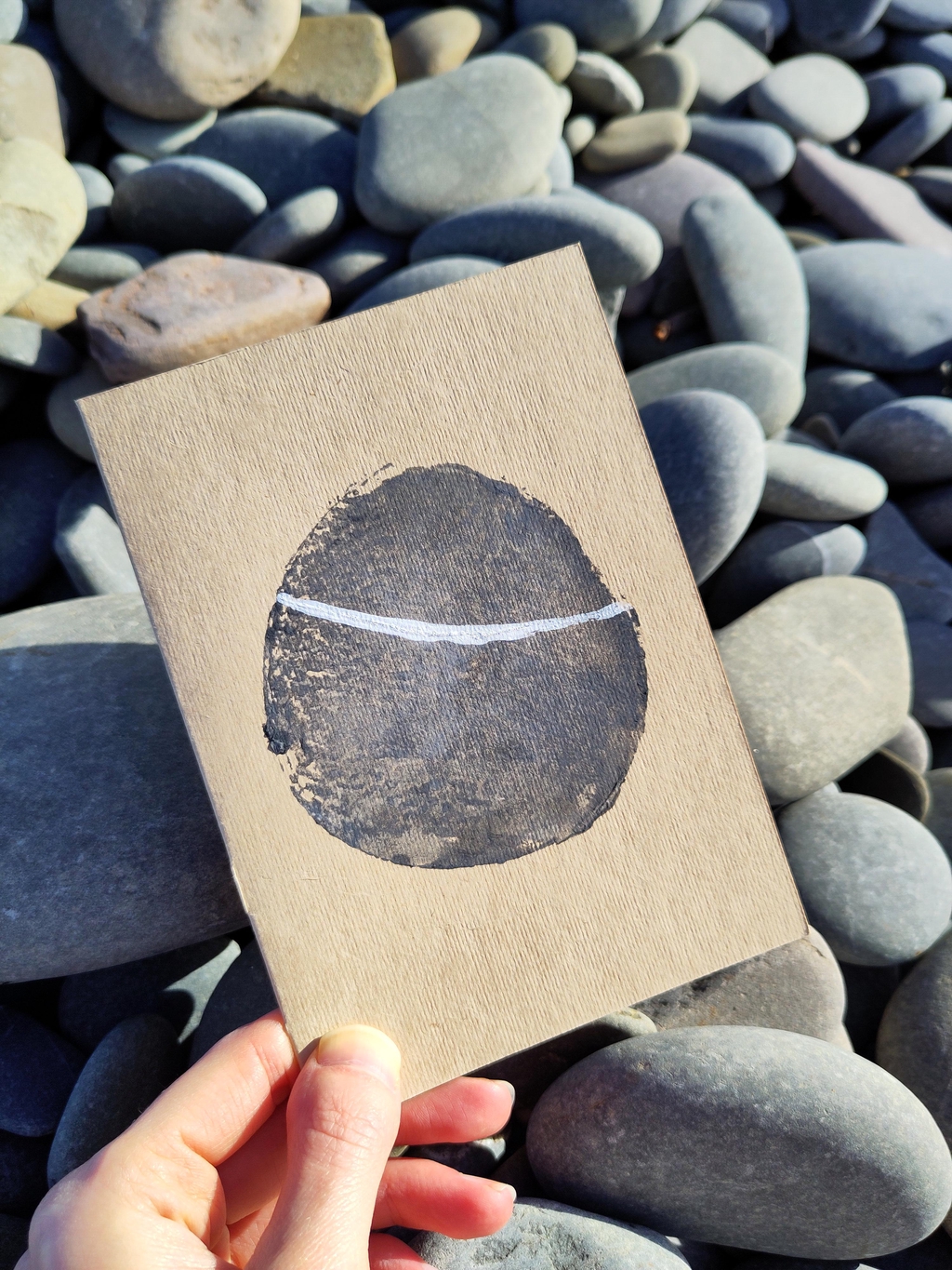A potato print of a smooth pebble on sandy brown card. There's a streak of white quartz running through the pebble. The card is resting on a pile of the real pebbles.
