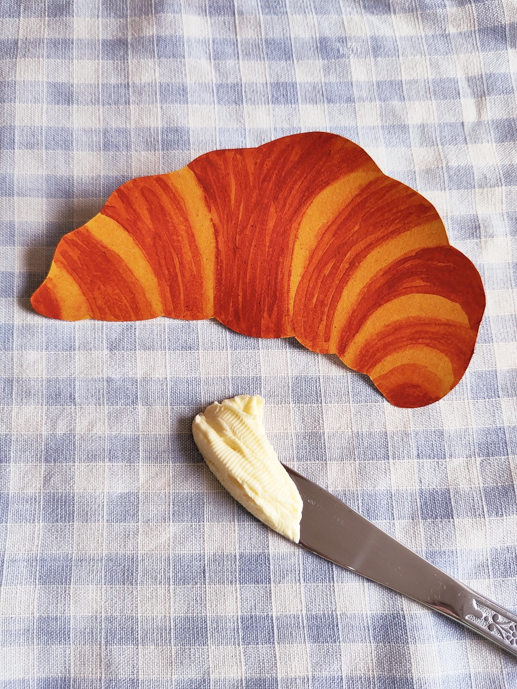 A cut-out watercolour of a croissant. It's resting on a light blue and white tablecloth next to a buttery knife.