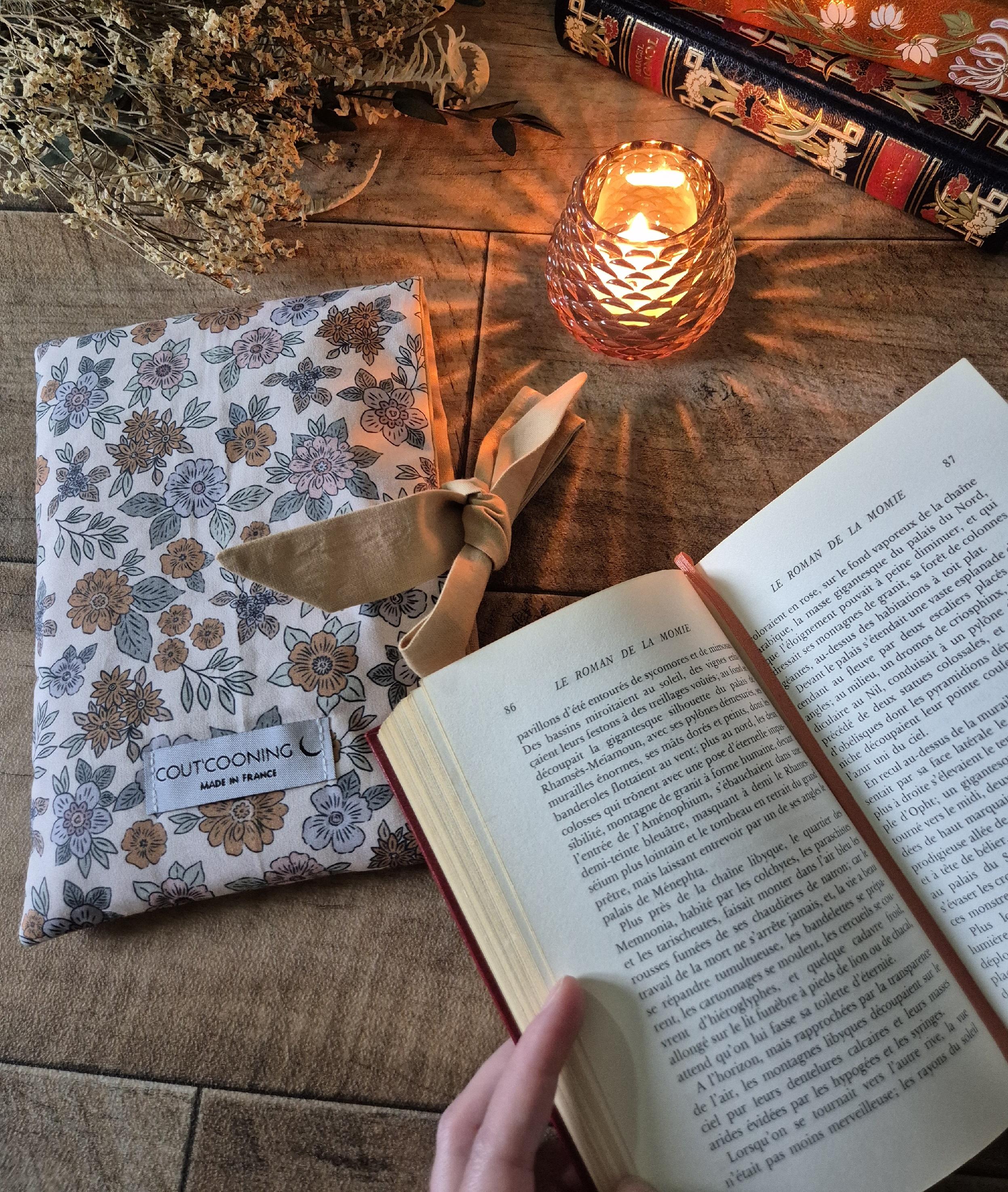 Pochette à livre artisanale cousue main mise en scène dans une ambiance douce et chaleureuse avec un livre ouvert et une bougie allumée Accessoire textile Amayë