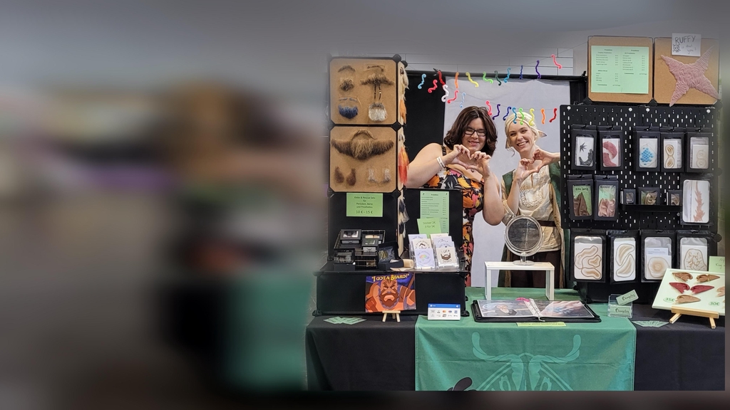 Messestand auf der Dokomi mit Bärten, Perücken und Silikonteilen. Zwei hellhäutige Frauen stehen dahinter und grüßen.