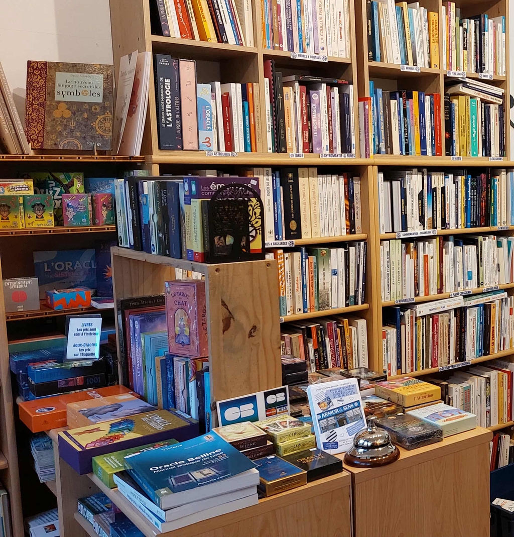 Présentation en boutique de livres d’occasion et de jeux d’oracles, exposés sur des étagères et un comptoir, avec panneaux de catégories et affichage des prix. 