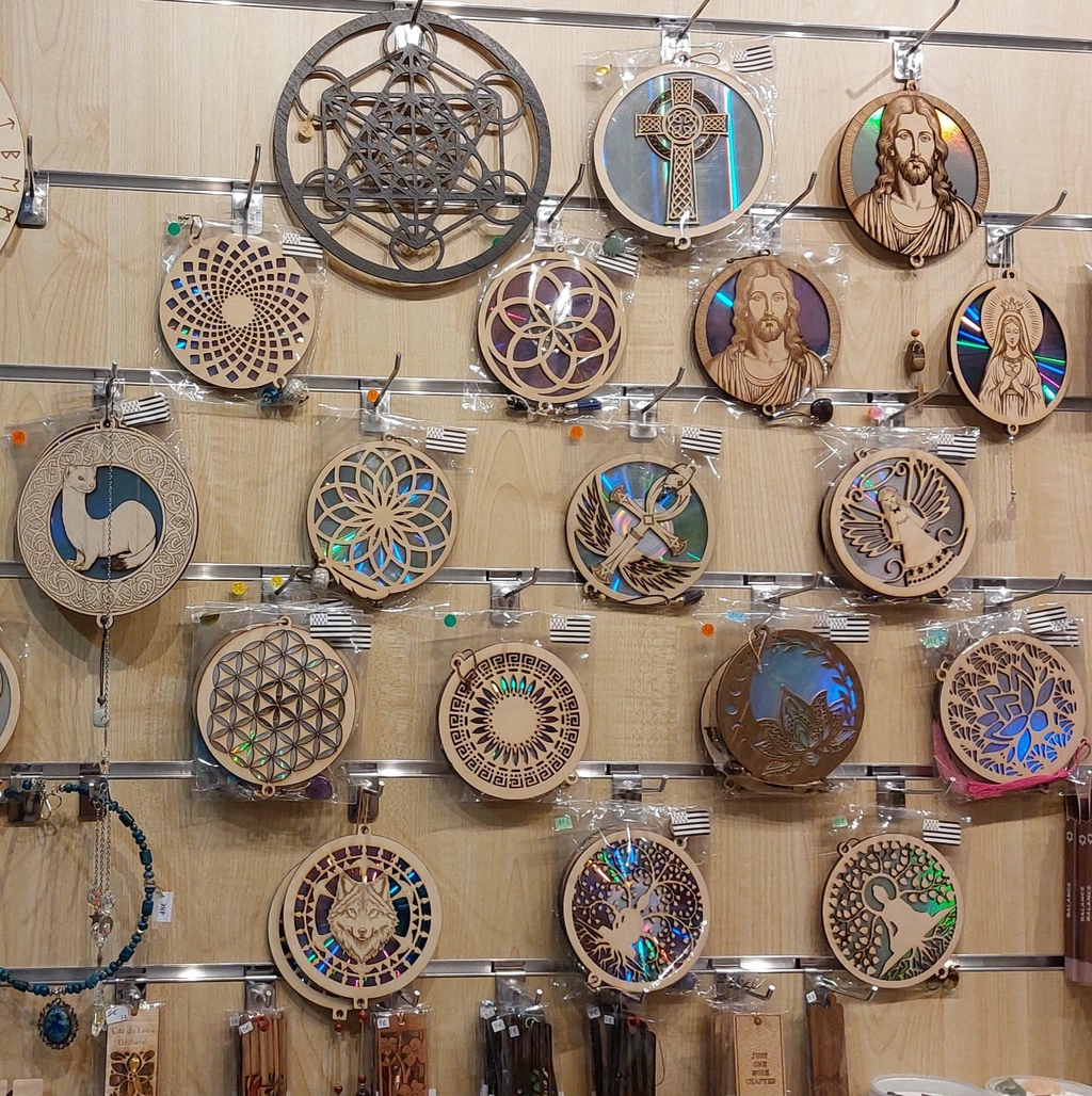 Présentation murale de décorations en bois gravé : motifs variés (animaux, symboles, mandalas, figures spirituelles) suspendus sur des crochets, avec porte-clés gravés, marque-pages gravés et/ou découpés, boucles d'oreilles gravés et/ou découpés