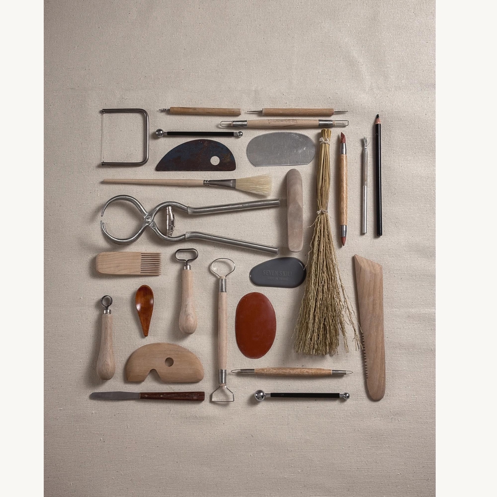 A curated flat lay of professional ceramic tools used by Rita Esteves: metal ribs, trimming loop tools, calipers, brushes, and wooden modeling tools on a linen background, representing the artisanal craft process.