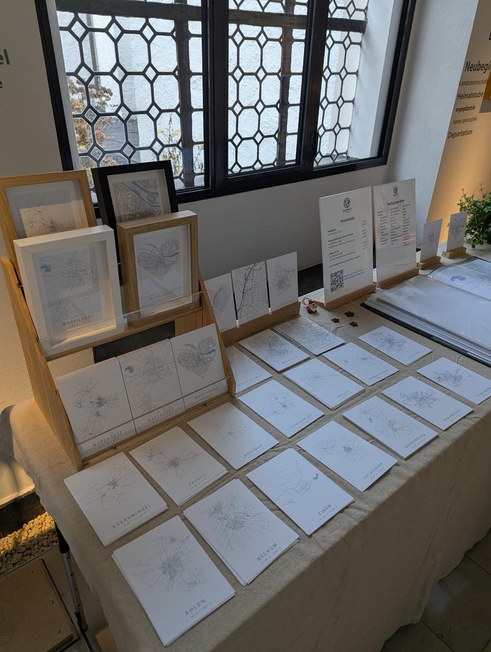Präsentation von lokalen Stadtkarten-Drucken auf einem Verkaufstisch: Links gerahmte Bilder im Holzaufsteller, mittig ausgelegte Postkarten verschiedener Städte und rechts Infotafeln zu Preisen und Verfügbarkeit.