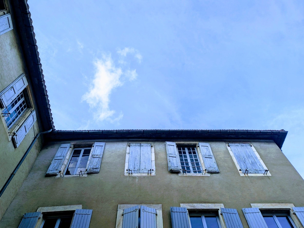 A view from the Maison de l'Aguze courtyard showing the blue sky, green trees, and blue shutters of the house. 